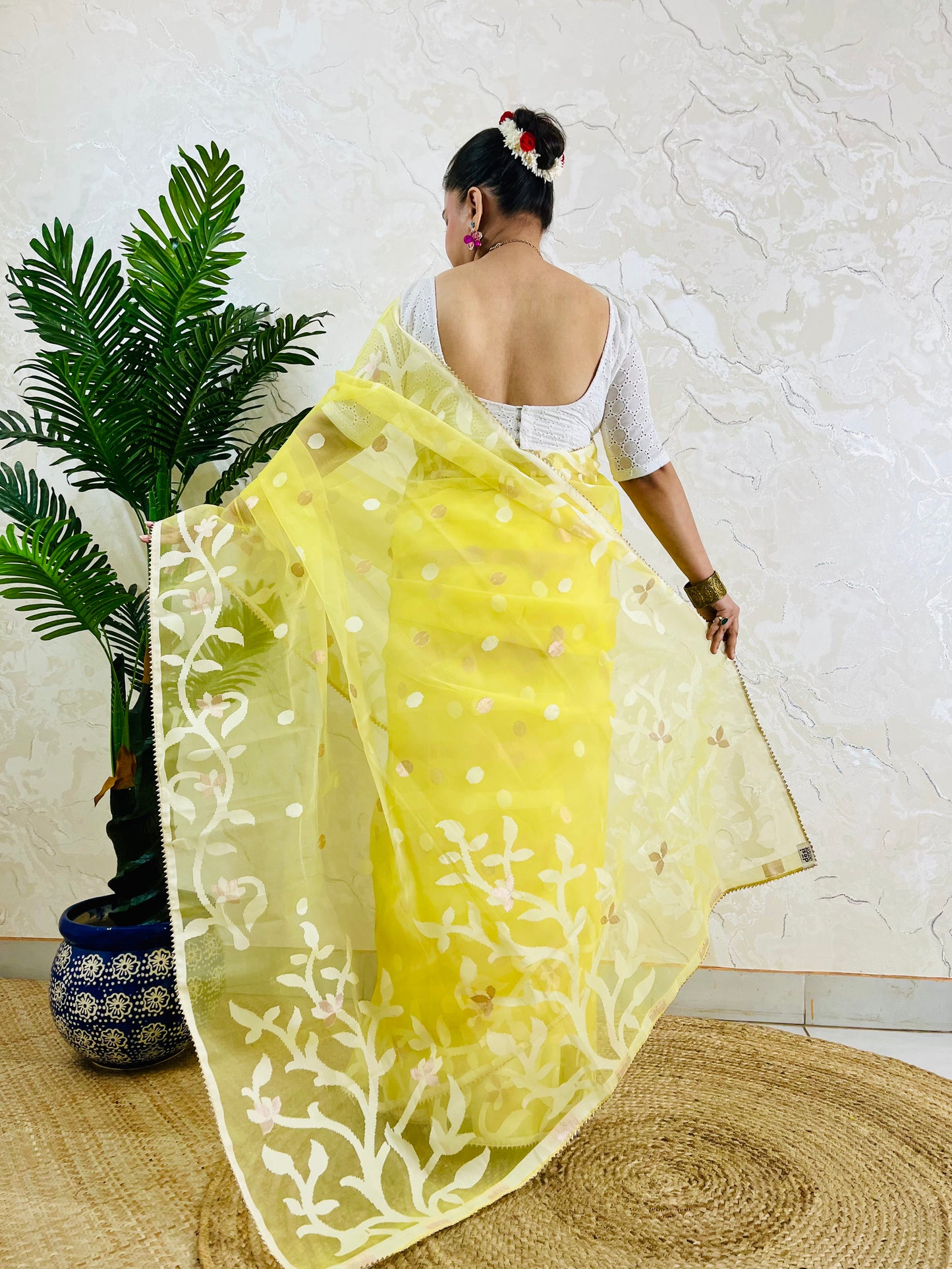 A woman wearing a bright yellow saree with white floral embroidery and a white sleeveless blouse, photographed from behind in an indoor setting with a potted plant and textured wall.