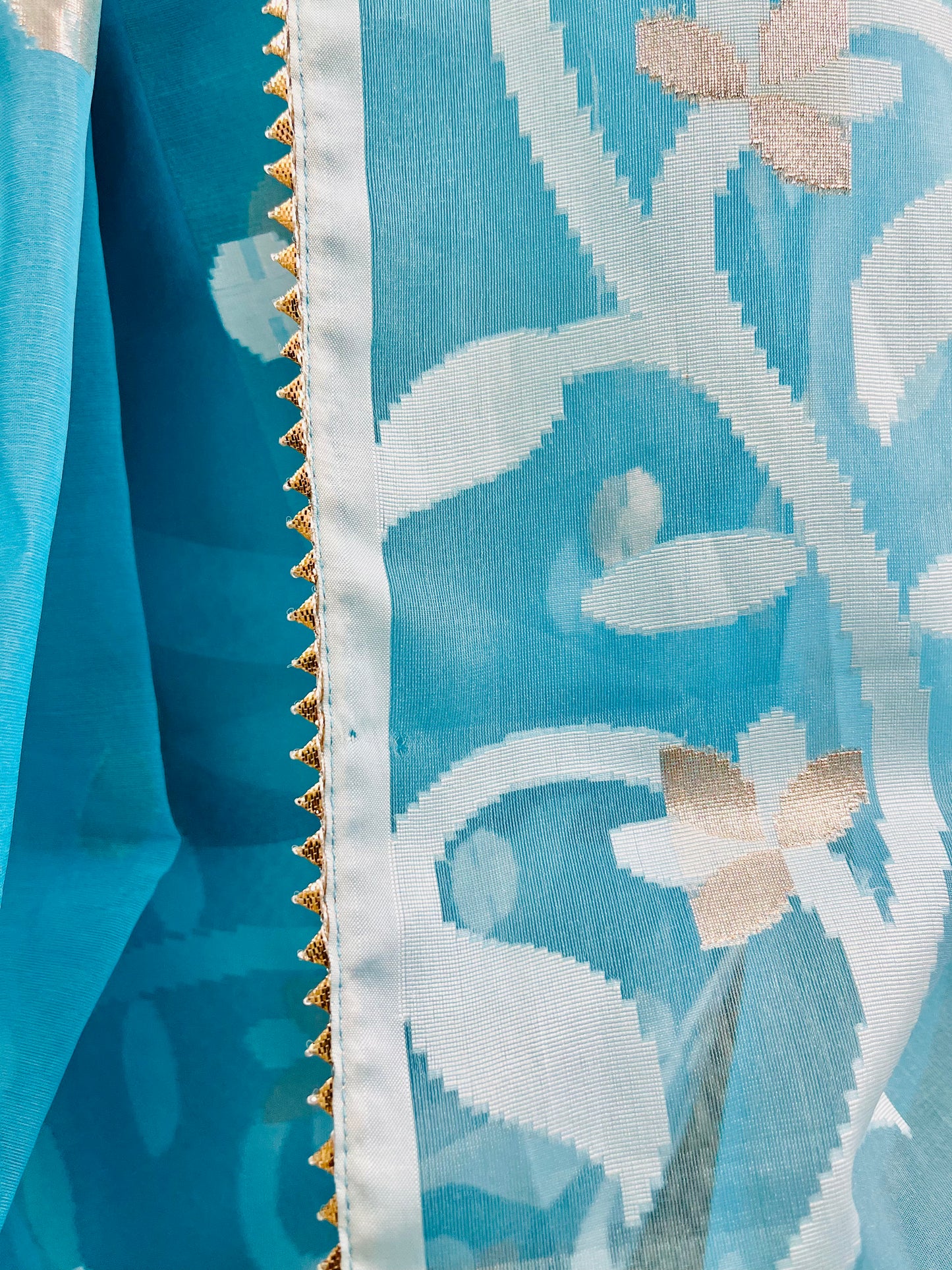 Close-up of a light blue sheer fabric with a white ikat-style floral pattern, gold border trim, and small beige flower accents.