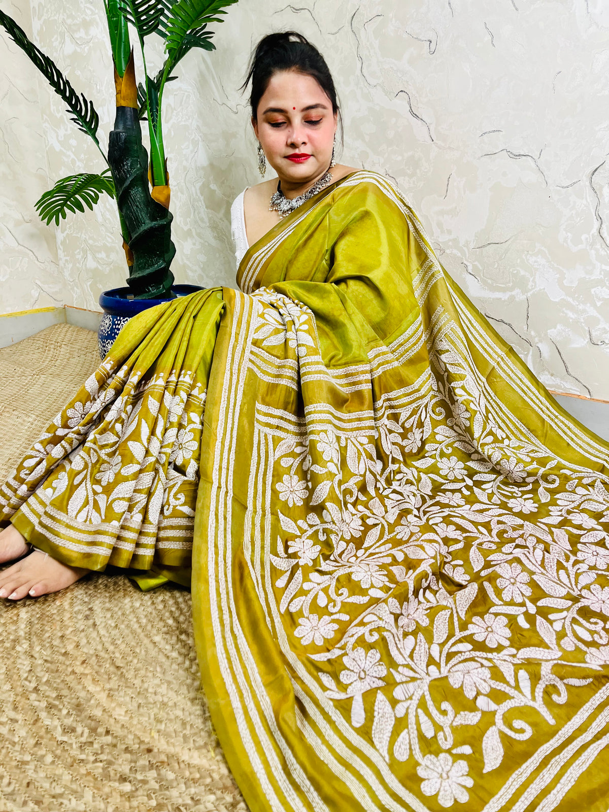 Woman sitting elegantly showcasing the dense, handcrafted white floral Katha embroidery on the pallu of the olive green Batik saree