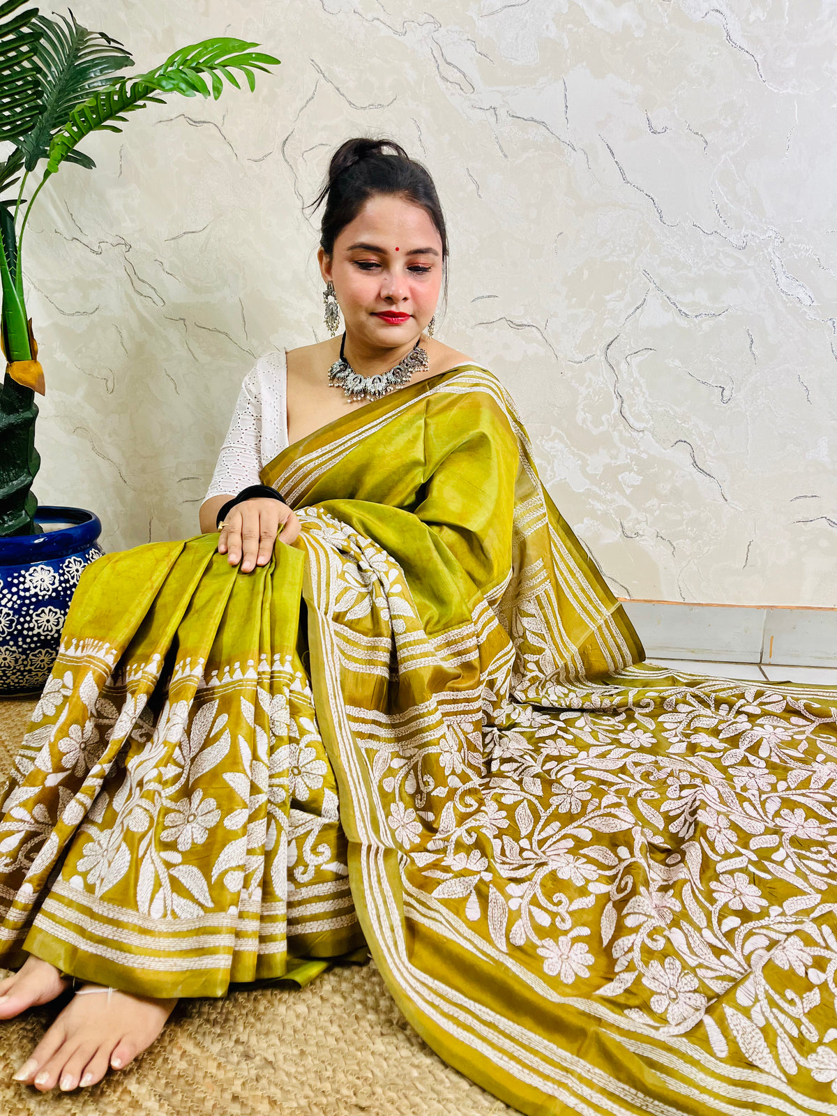 Woman sitting elegantly showcasing the dense, handcrafted white floral Katha embroidery on the pallu of the olive green Batik saree