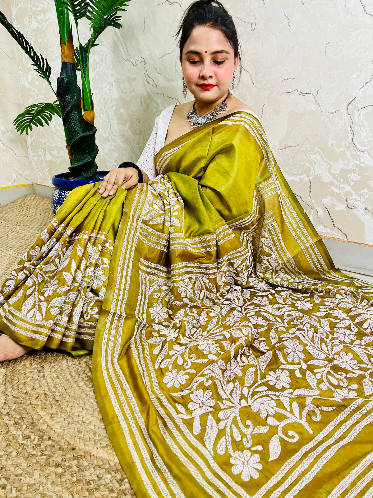 Woman sitting elegantly showcasing the dense, handcrafted white floral Katha embroidery on the pallu of the olive green Batik saree