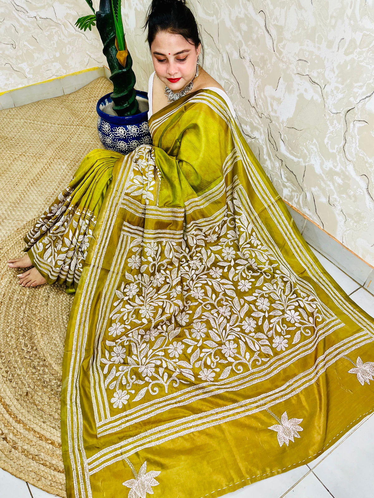 Woman sitting elegantly showcasing the dense, handcrafted white floral Katha embroidery on the pallu of the olive green Batik saree