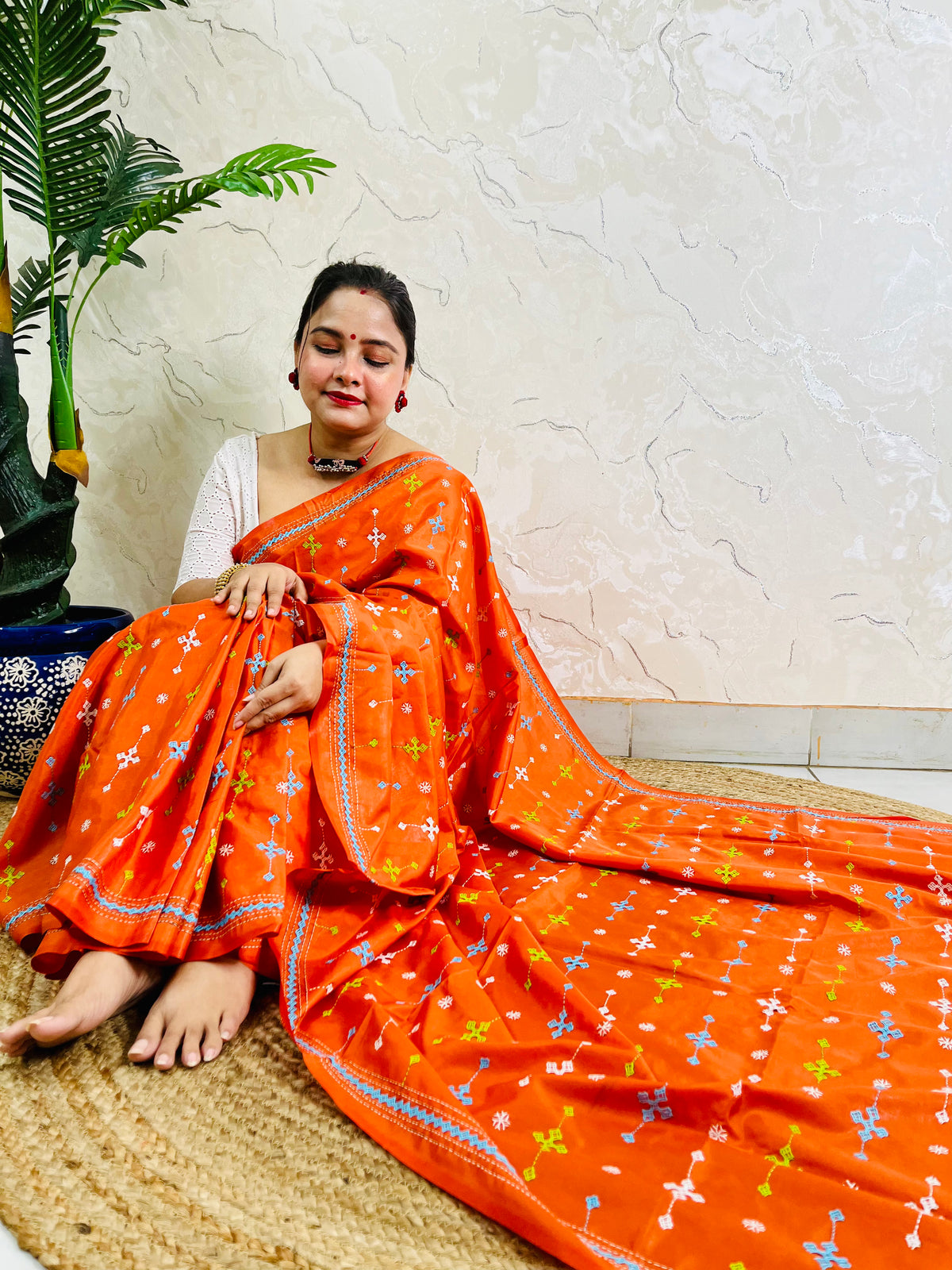 Woman seated in orange saree with blue and green cross-stitch embroidery detail
