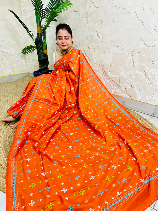 Woman seated in orange saree with blue and green cross-stitch embroidery detail