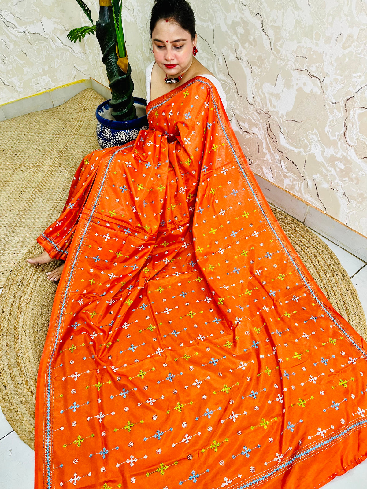 Woman seated in orange saree with blue and green cross-stitch embroidery detail