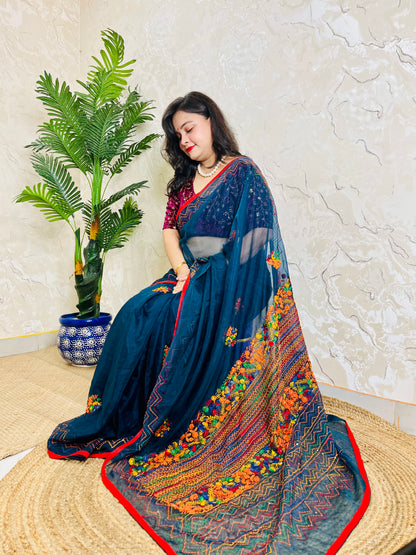 Woman wearing Desi Aadat handcrafted indigo organza silk saree with bullion stitch floral border and French knot embroidery, seated in an elegant pose — luxury ethnic wear for festive occasions