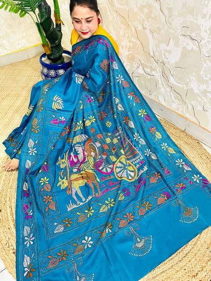 Woman seated in peacock blue Kantha saree, side pallu spread showing bullock cart scene