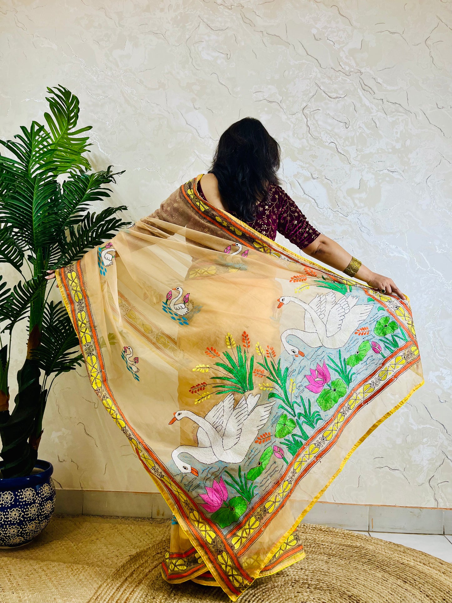 Back standing view of beige-gold organza saree with Kantha stitch swan and lotus embroidery, pallu flowing