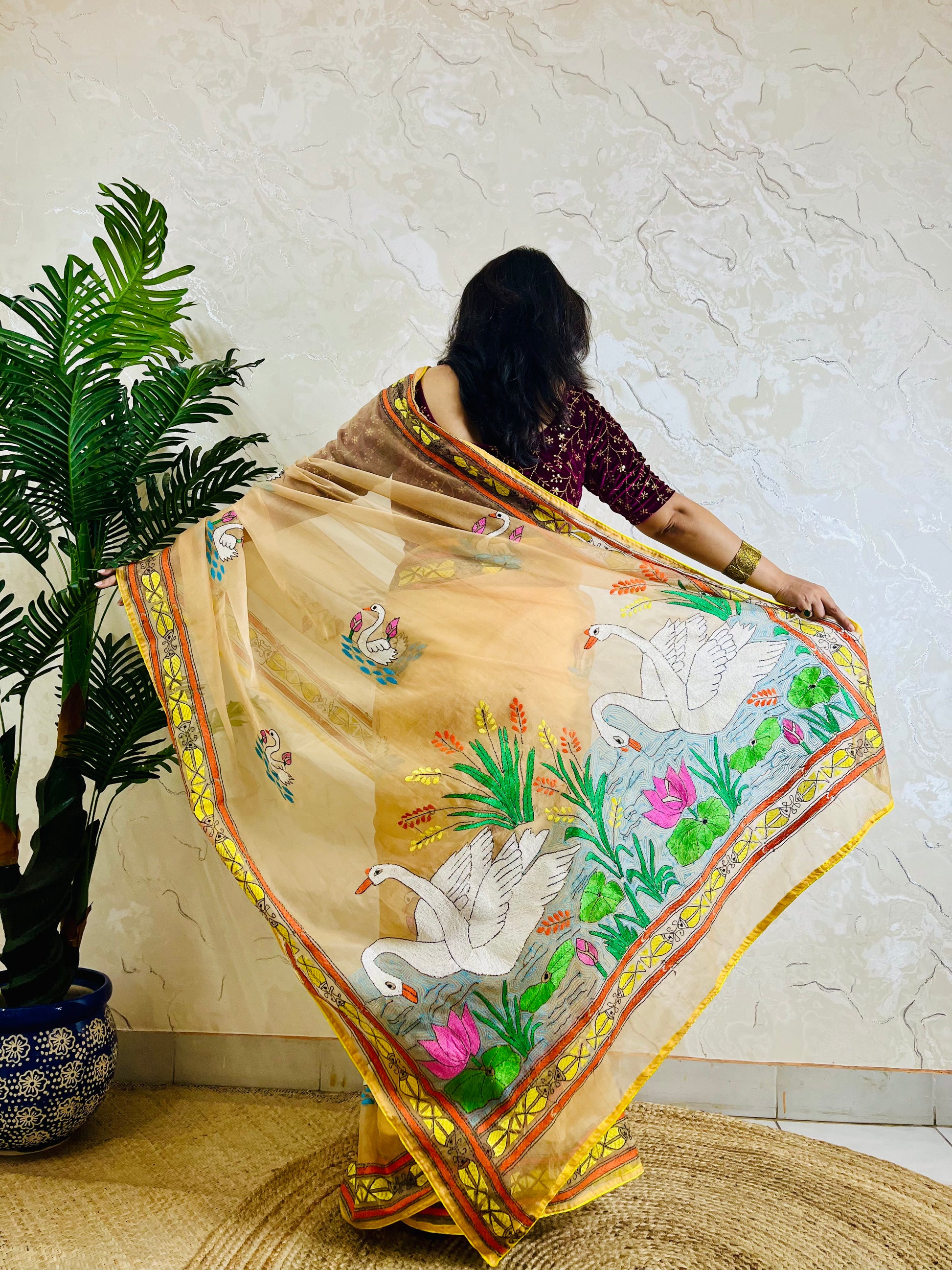 Back standing view of beige-gold organza saree with Kantha stitch swan and lotus embroidery, pallu flowing