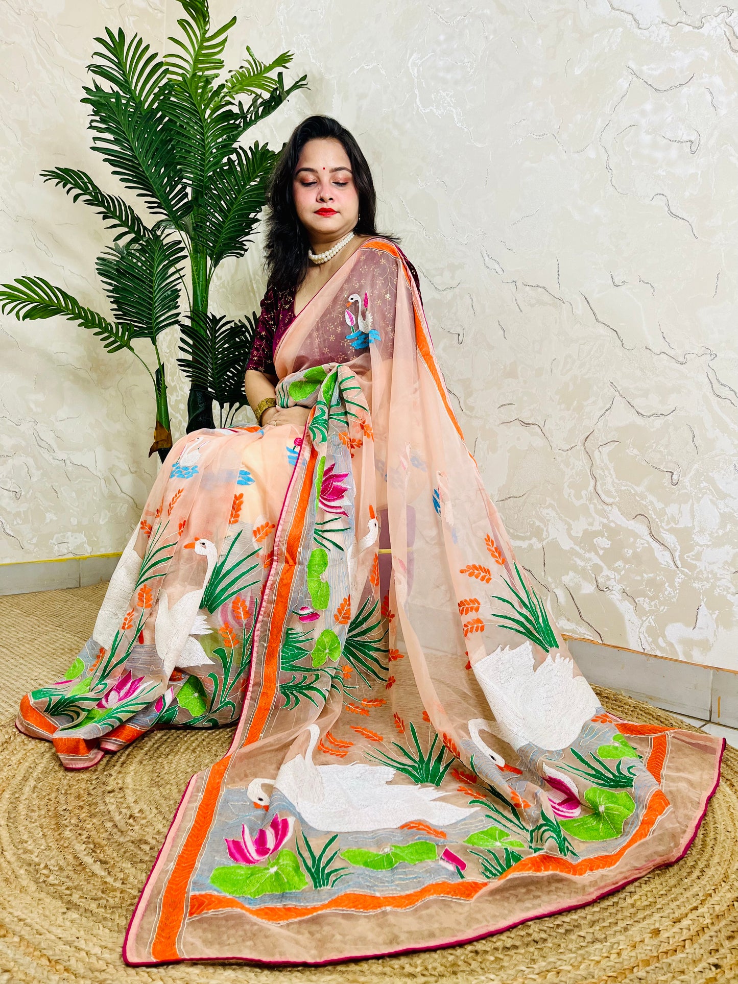 Woman seated in peach-pink Kantha stitch organza saree with swan and lotus embroidery, pallu draped in front