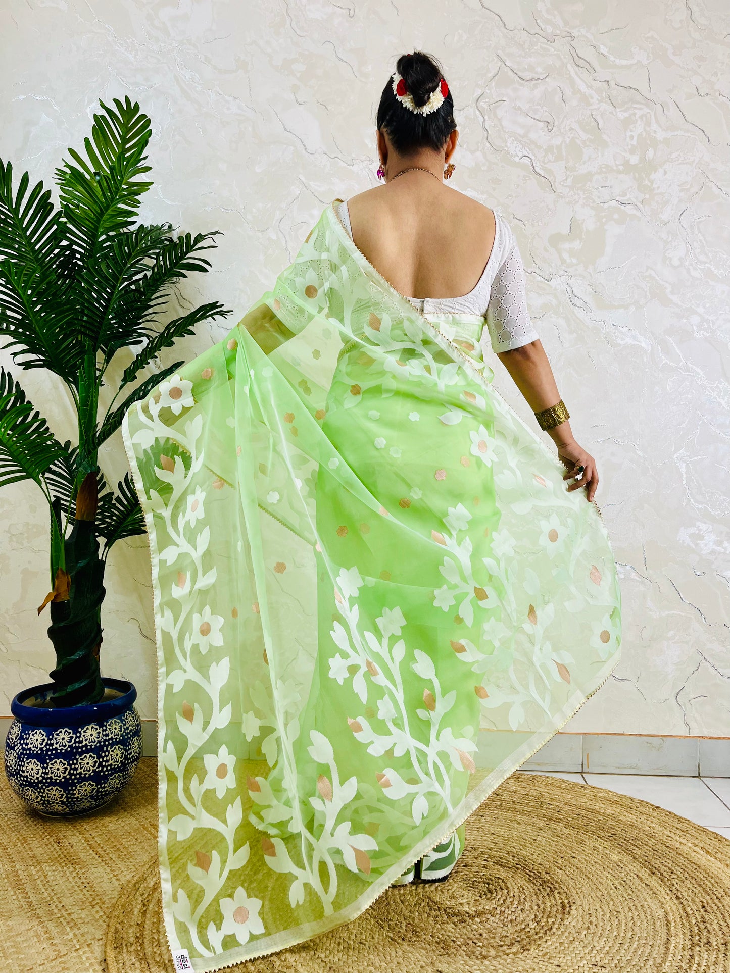 A woman viewed from behind wearing a light green saree with white floral embroidery and small gold motifs, paired with a white blouse. She is standing on a round woven mat in front of a textured beige wall, with a potted green plant on the left.
