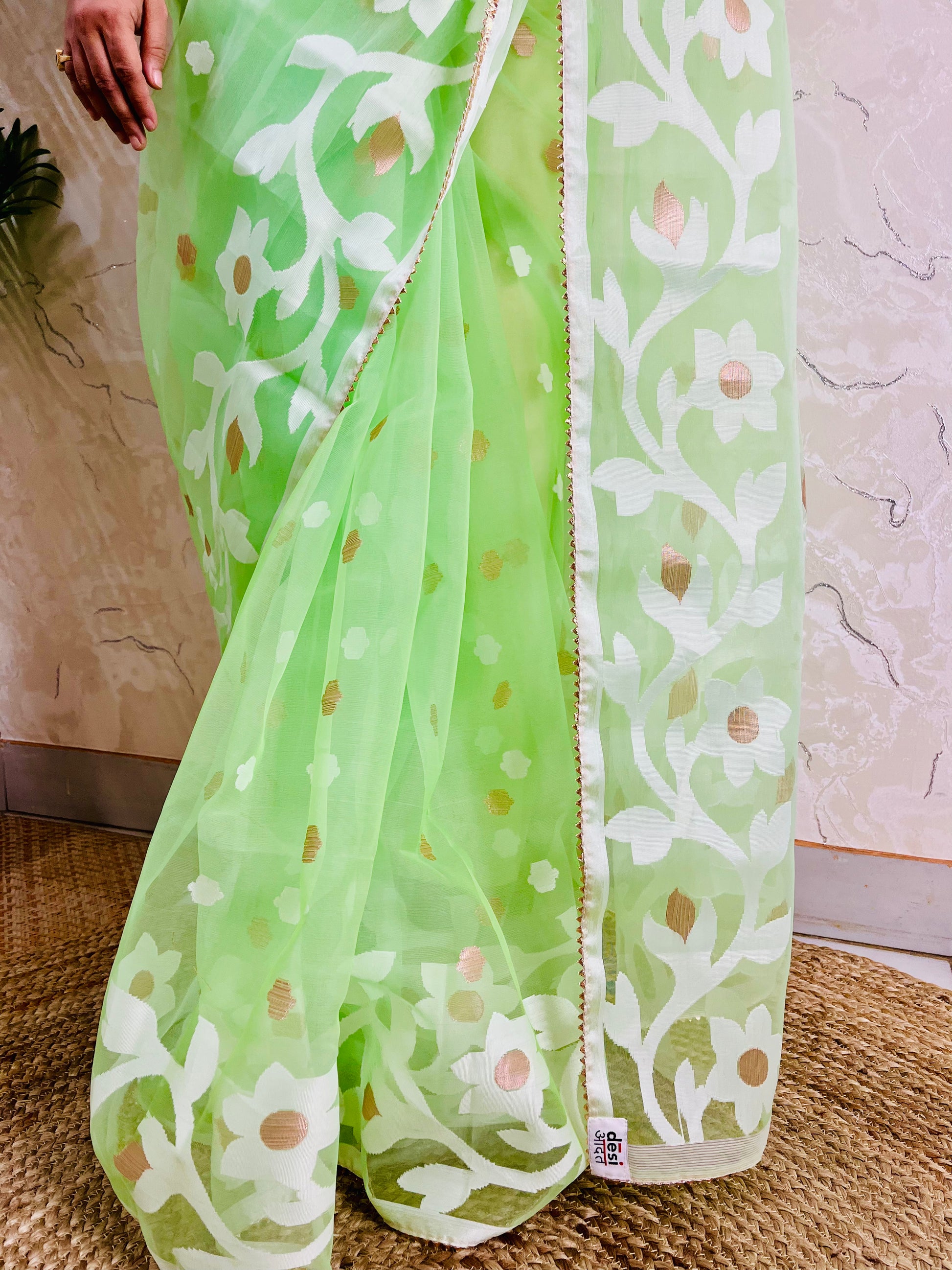 A light green saree with white floral patterns and small gold foil accents, draped on a model. The image shows the lower half of the model and the drape of the saree, set against a neutral indoor background.
