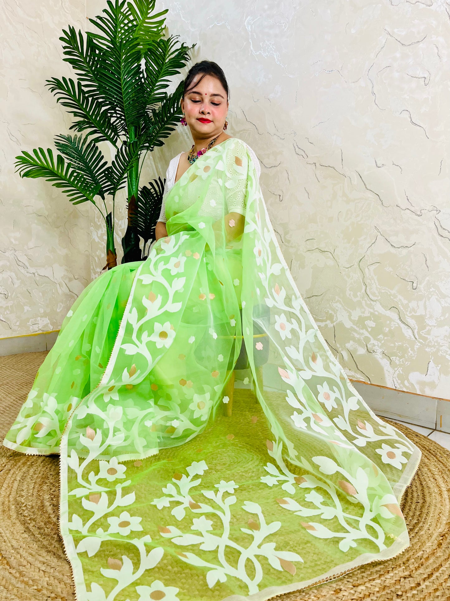 A woman wearing a bright lime green saree with white floral embroidery, seated on a woven mat in front of a textured beige wall and a potted plant.