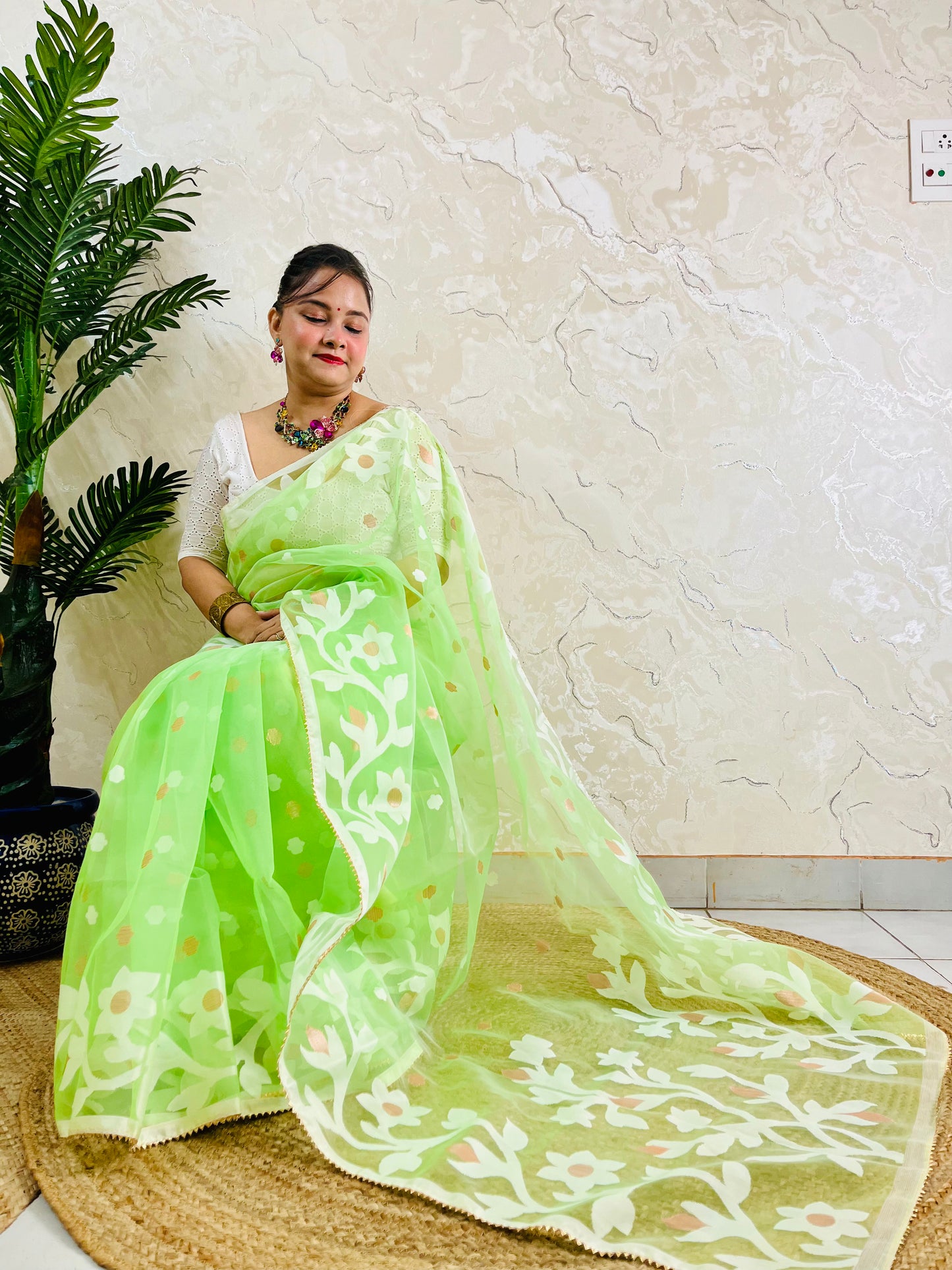 A woman wearing a light green saree with white floral embroidery and a white blouse, seated on a woven mat against a textured beige wall with a potted plant on the left.