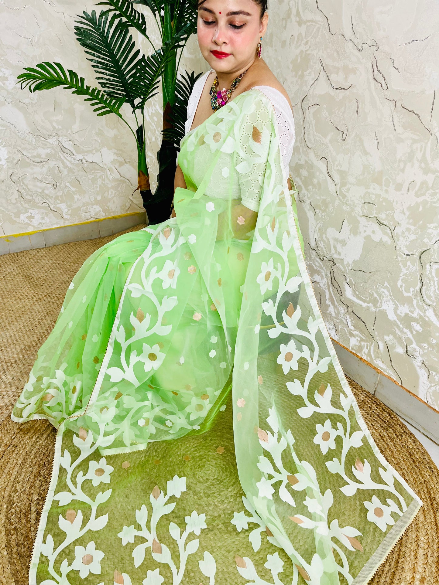 A woman wearing a light green embroidered saree with white floral patterns and a matching dupatta, seated on a woven mat against a textured wall and potted plant.