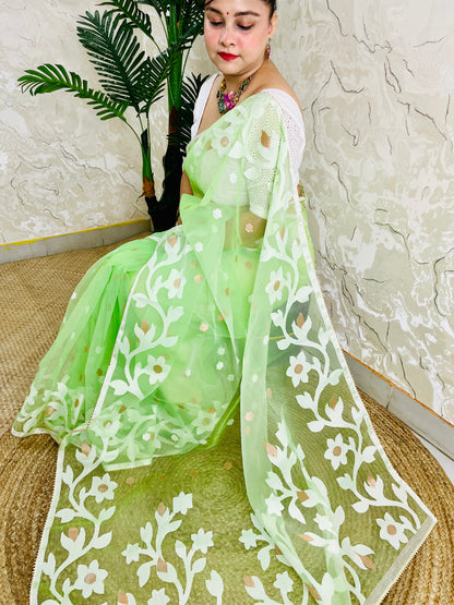 A woman wearing a light green embroidered saree with white floral patterns and a matching dupatta, seated on a woven mat against a textured wall and potted plant.