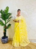 A woman wearing a bright yellow embroidered saree with a white blouse stands indoors against a textured cream wall, next to a potted green plant.