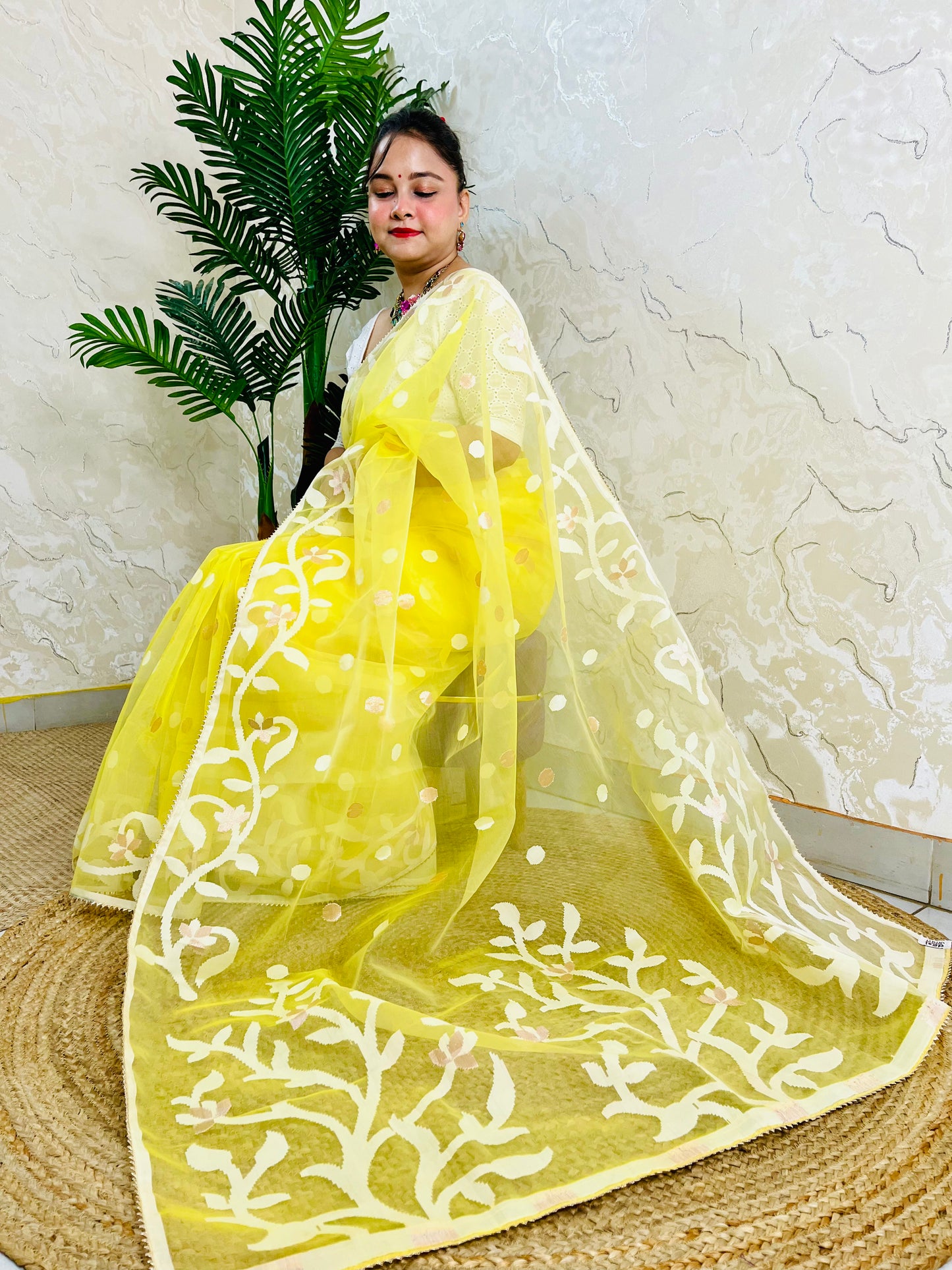 A woman wearing a bright yellow traditional outfit with a sheer embroidered dupatta draped over a woven mat, posed in front of a textured wall and a potted plant.