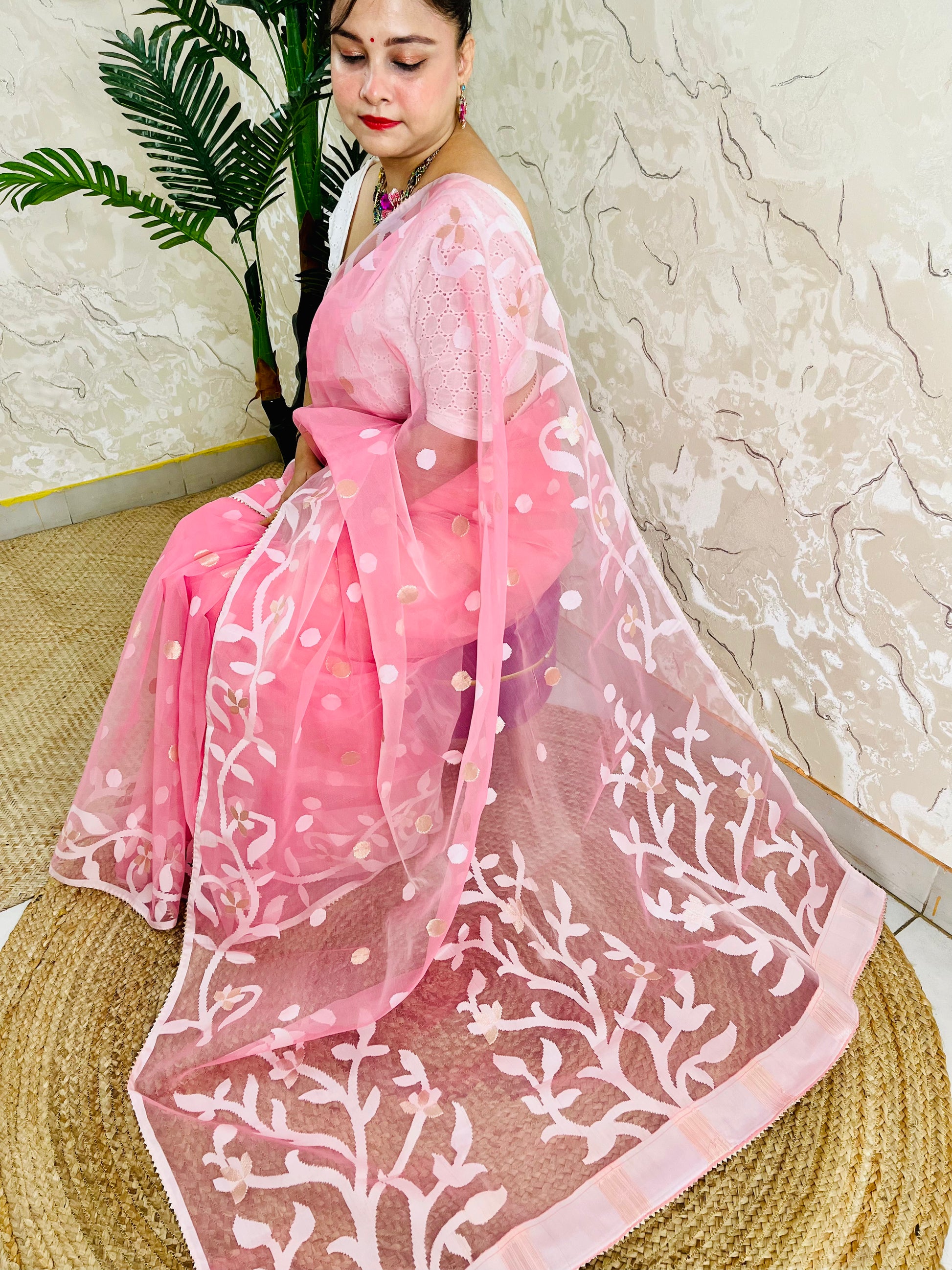 A woman seated indoors wearing a pink saree with a sheer pink dupatta featuring white floral embroidery and small gold motifs, set against a textured wall and woven mat.