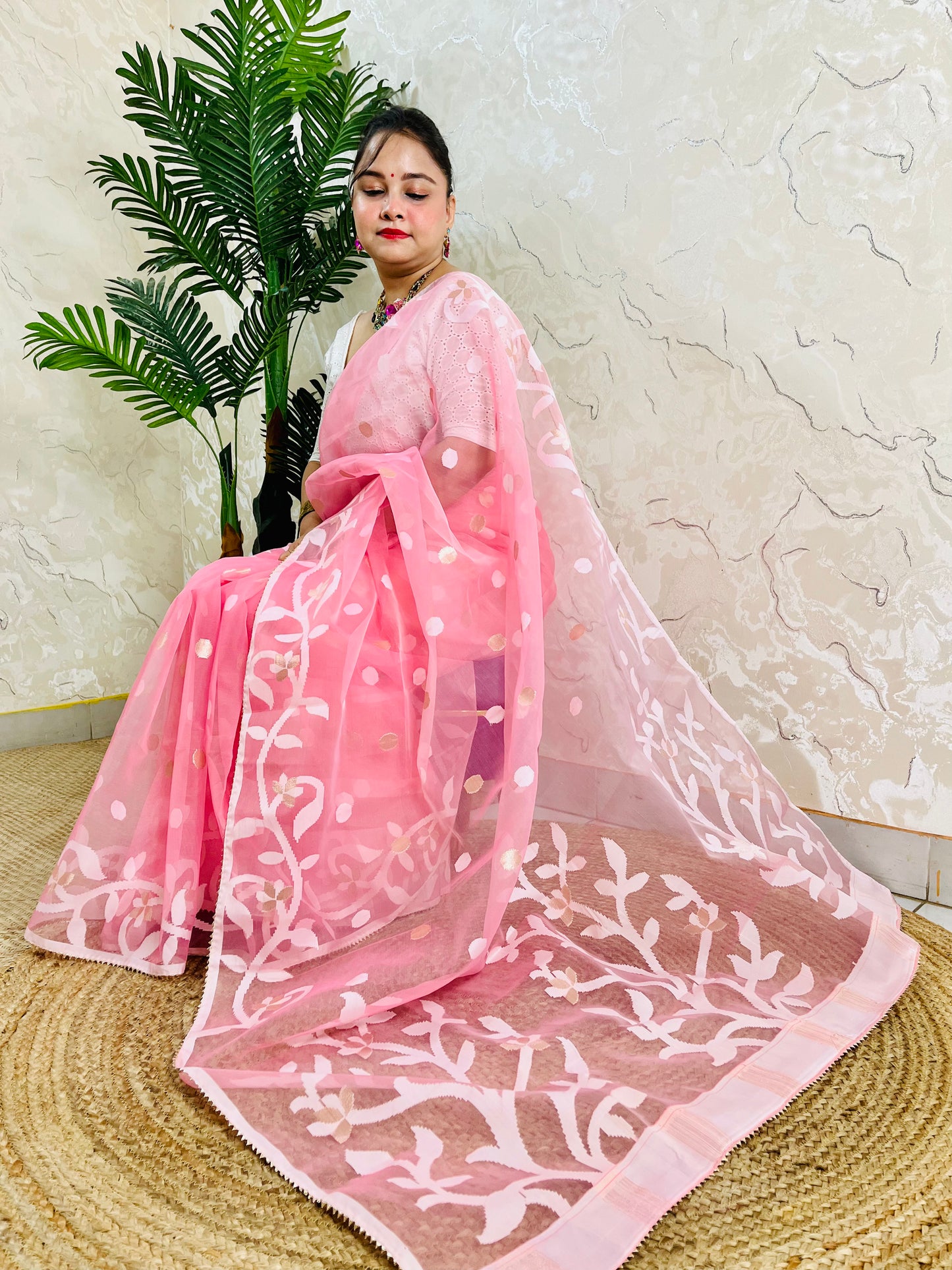A woman wearing a pink saree with white floral embroidery and gold polka dots, posed in front of a textured beige wall with a green plant in the background.