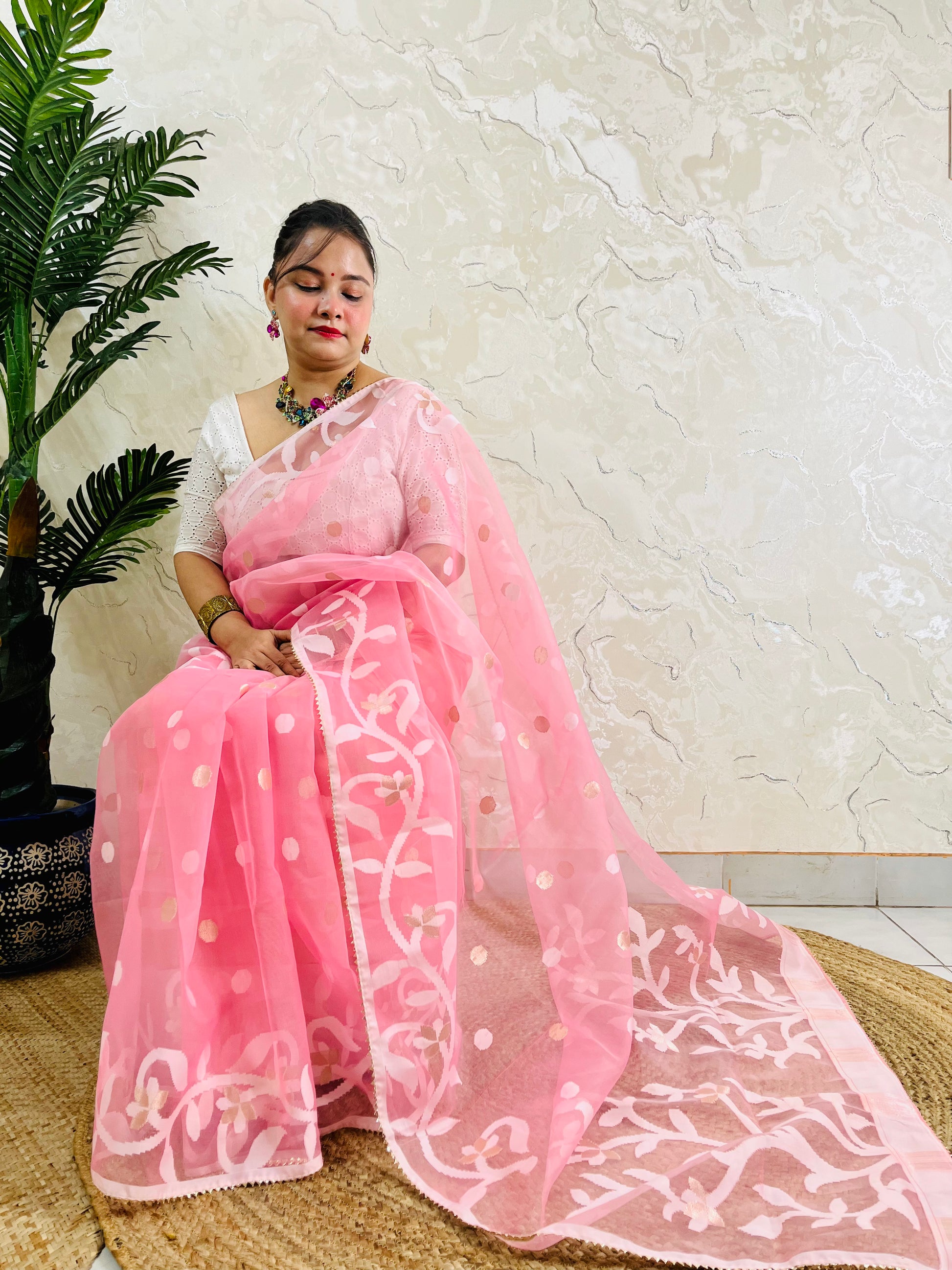 A woman wearing a light pink saree with white floral embroidery and gold polka dots, paired with a white sleeveless blouse, sitting on a woven mat against a textured beige wall.