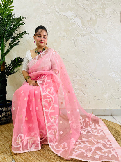 A woman wearing a light pink saree with white floral embroidery and gold polka dots, paired with a white sleeveless blouse, sitting on a woven mat against a textured beige wall.