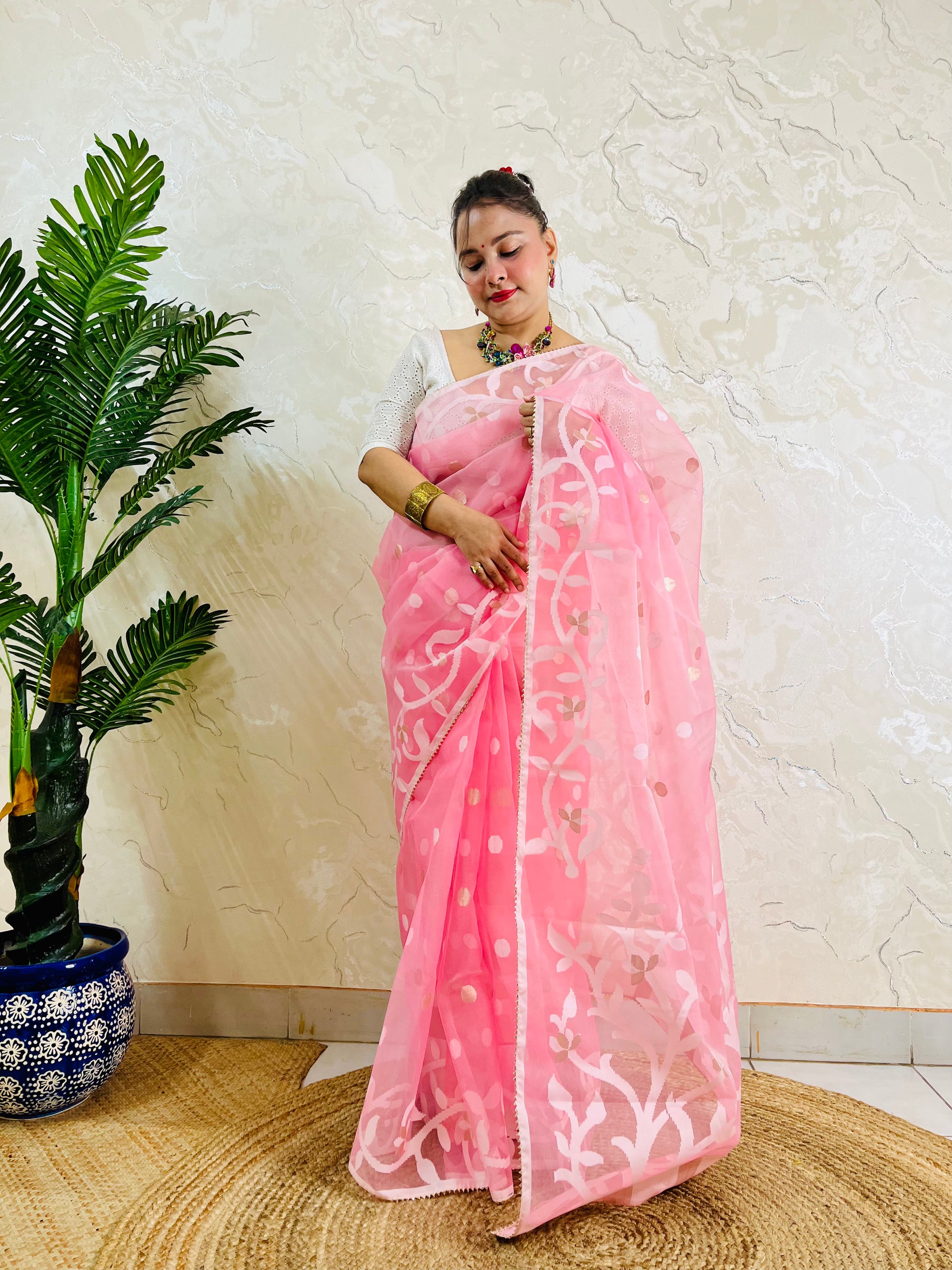 A woman wearing a pink saree with white floral patterns and a white blouse, standing indoors against a textured beige wall next to a potted green plant.