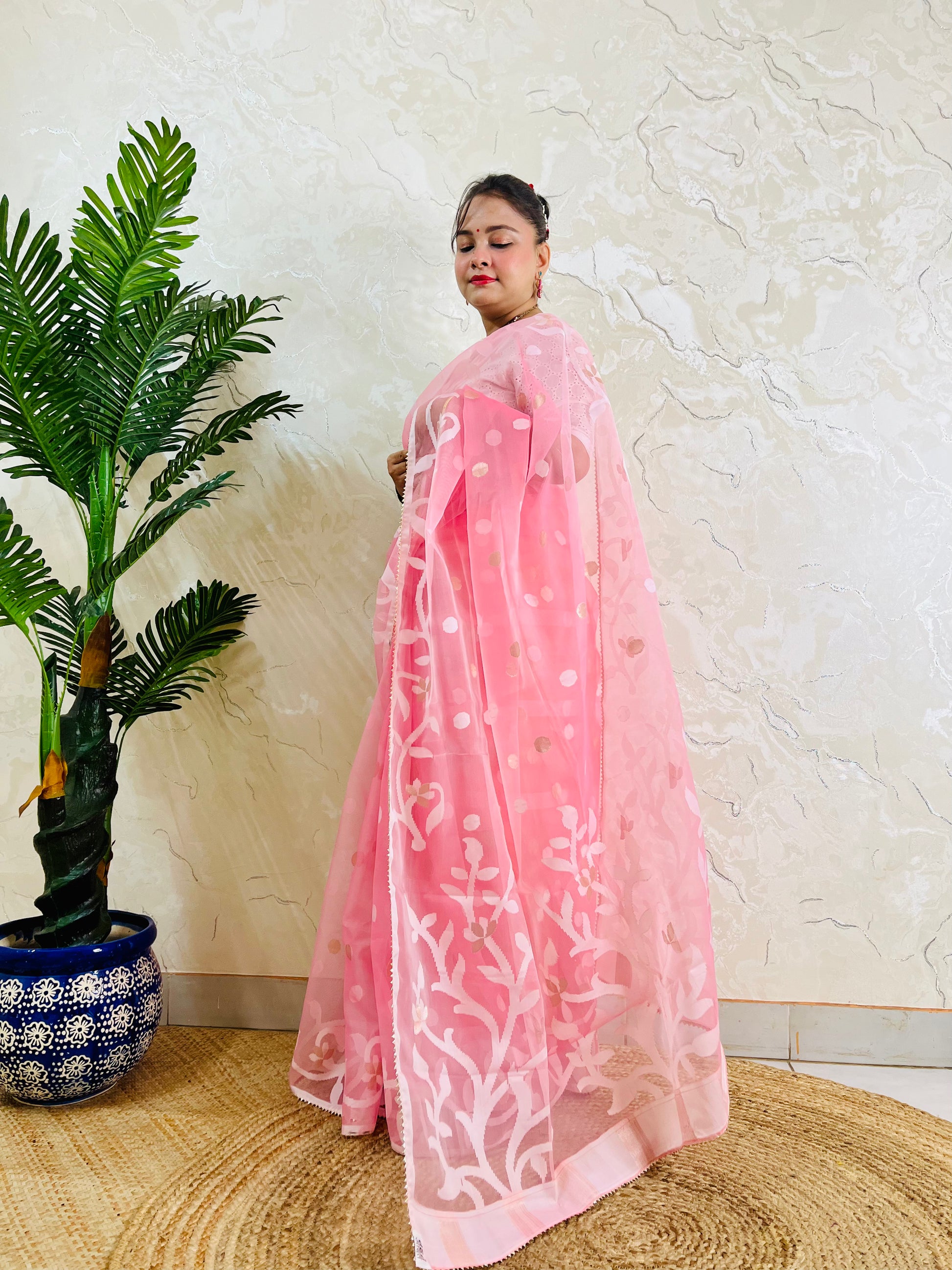 A woman wearing a pink organza saree with white floral embroidery, standing in a room with a textured beige wall and a potted green plant on the left.