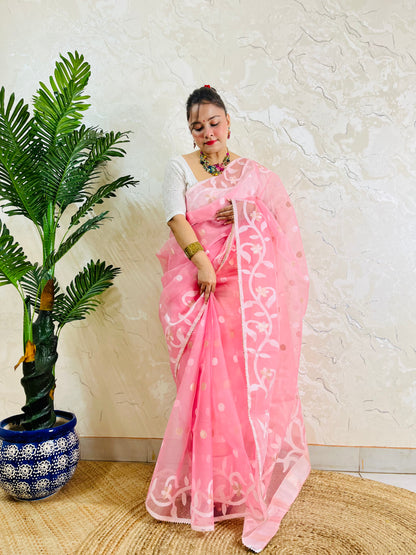 A woman wearing a pink saree with white floral embroidery and gold polka dots, paired with a white sleeveless blouse, standing next to a potted palm tree against a textured beige wall.