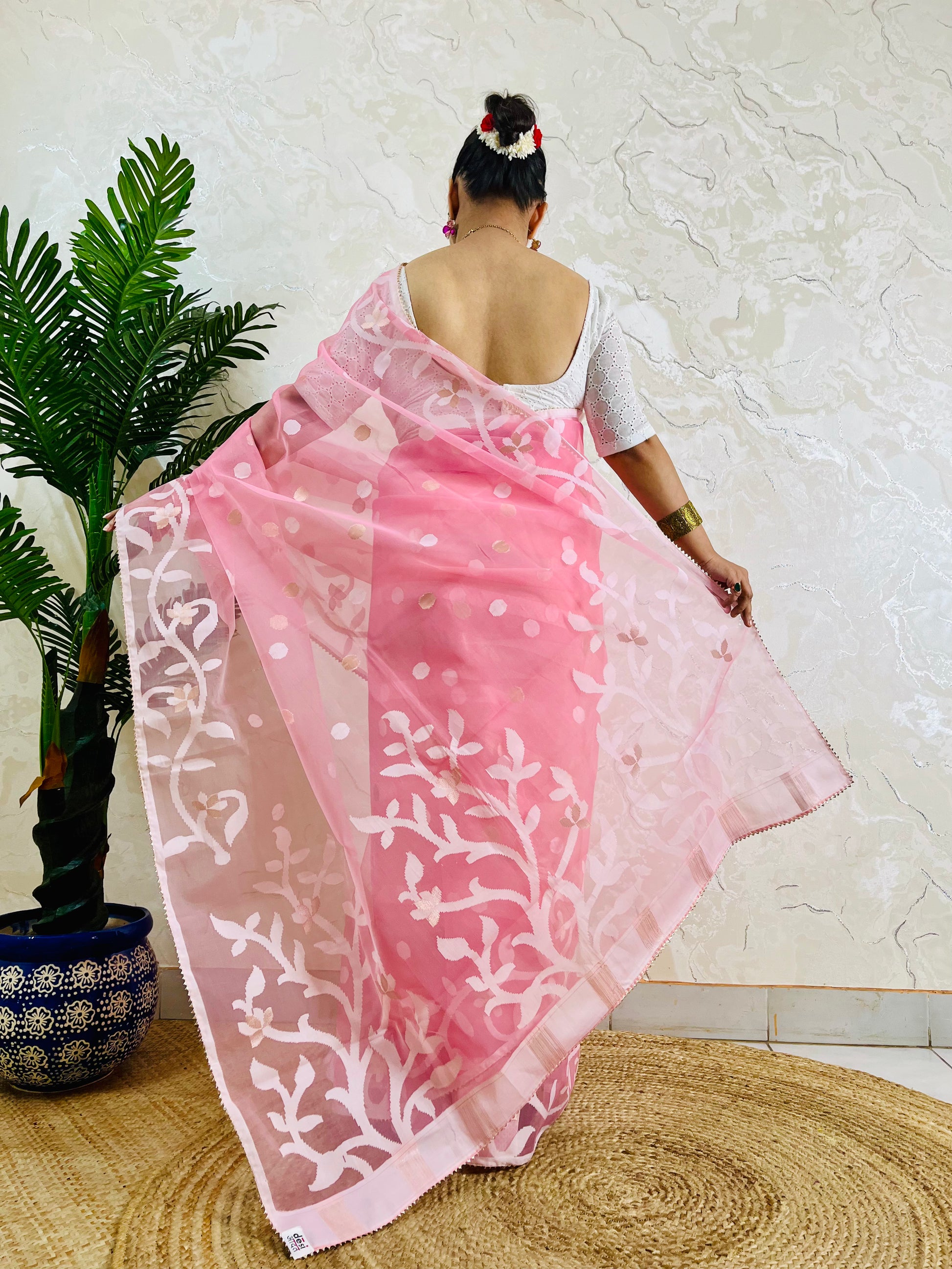 A woman viewed from behind wearing a pink saree with white floral and dot patterns, holding it out to show the fabric design. She is standing indoors against a textured off-white wall on a woven mat, next to a potted green plant.