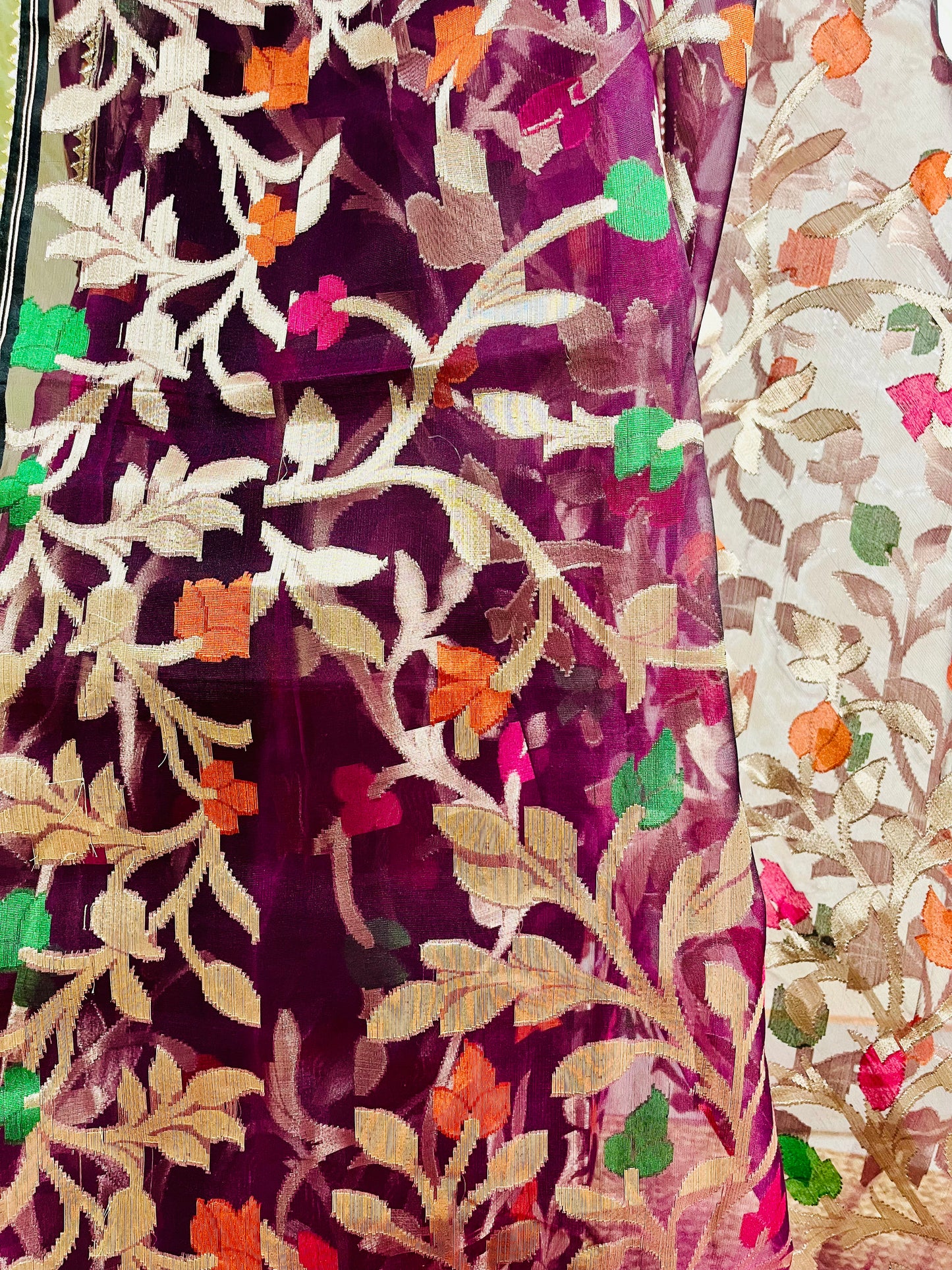 Close-up of a sheer magenta fabric with intricate floral embroidery in beige, orange, green, and pink, alongside a cream-colored embroidered fabric panel.