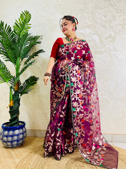 A woman models a purple and magenta floral embroidered saree with a red blouse, standing next to a potted palm plant against a textured cream wall.