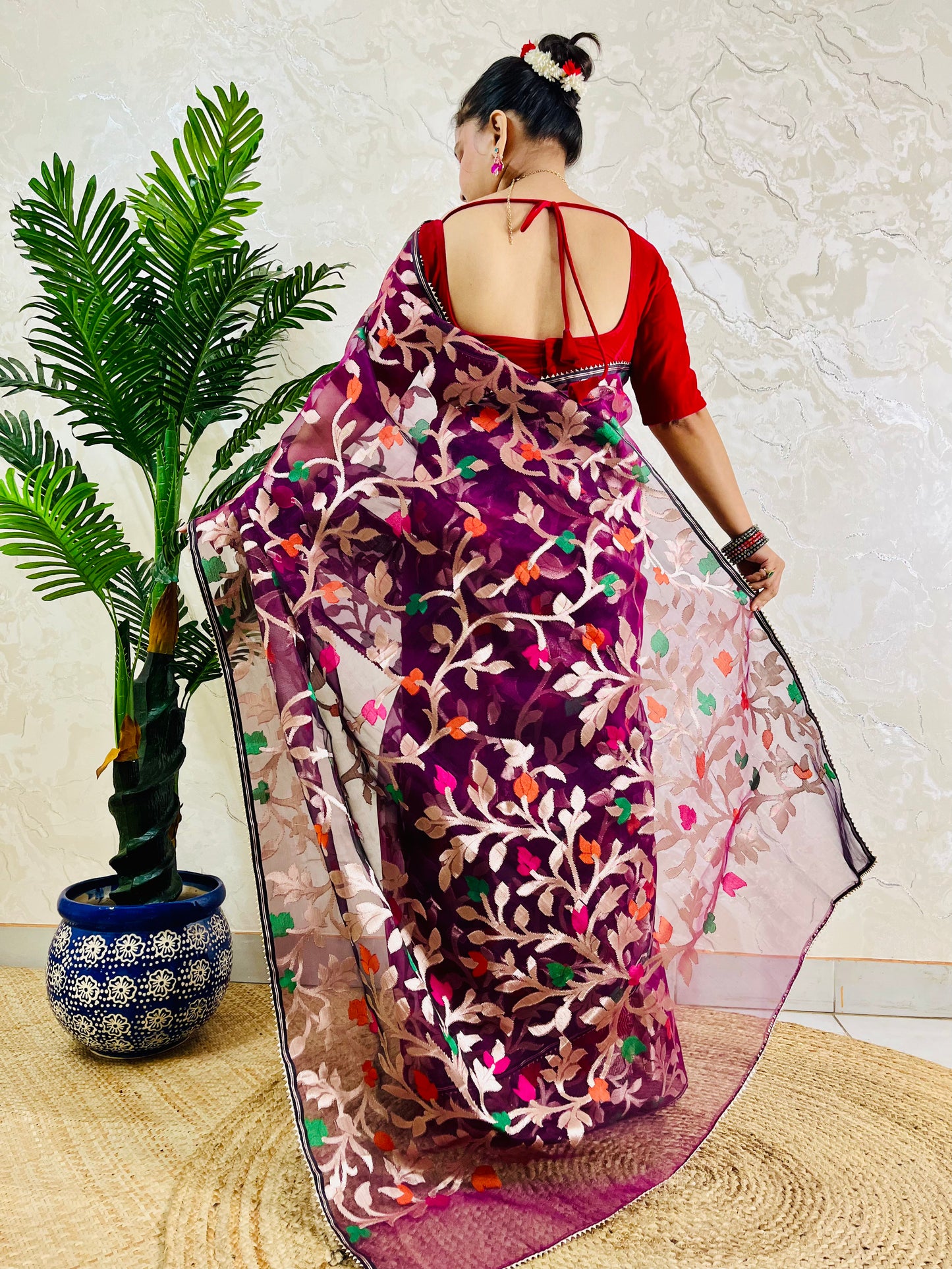A woman stands with her back to the camera wearing a purple and pink saree with intricate floral embroidery in pink, orange, and green, paired with a red blouse. She is standing on a woven mat next to a potted palm tree against a textured beige wall.