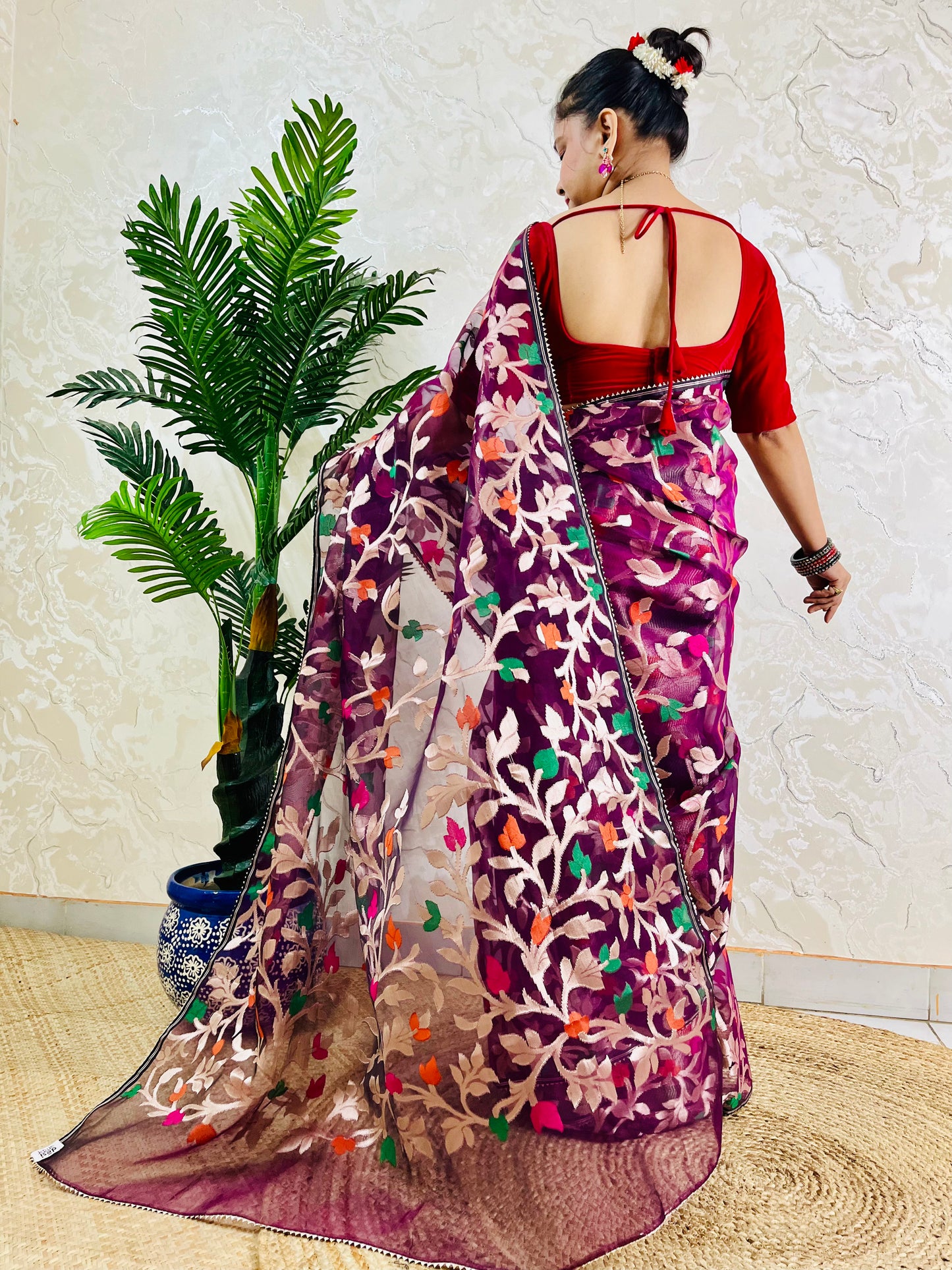 A woman wearing a purple and magenta floral embroidered saree with a red blouse, shown from the back in a home setting with a potted palm plant.