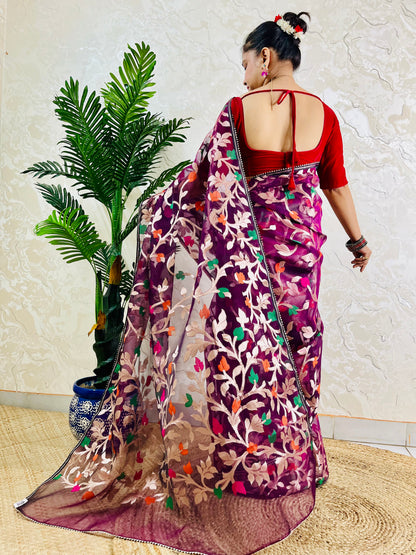 A woman wearing a purple and magenta floral embroidered saree with a red blouse, shown from the back in a home setting with a potted palm plant.