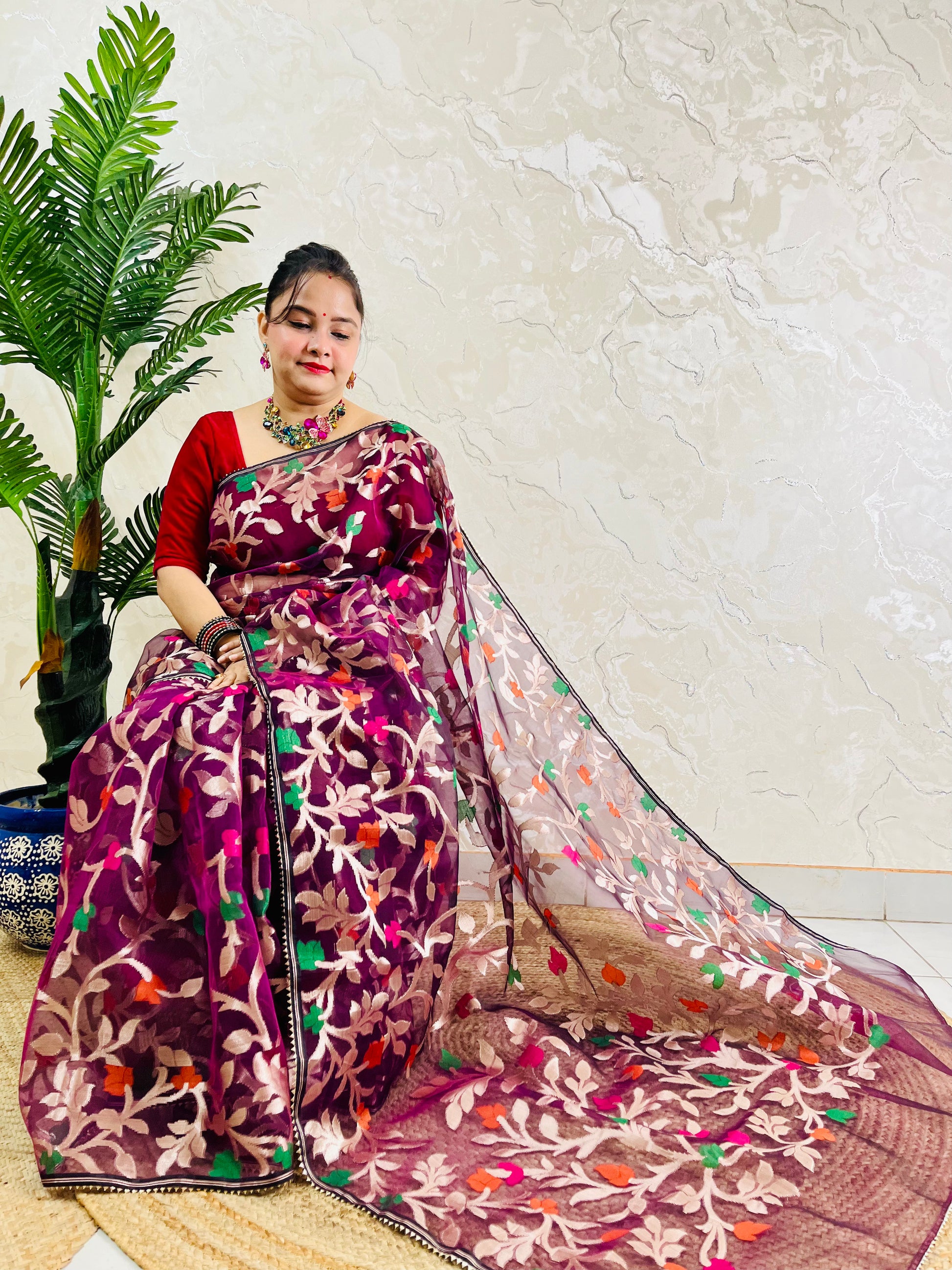 A woman wearing a purple and pink floral embroidered saree with a red blouse, seated on a woven mat against a textured beige wall with a green potted plant to the side.
