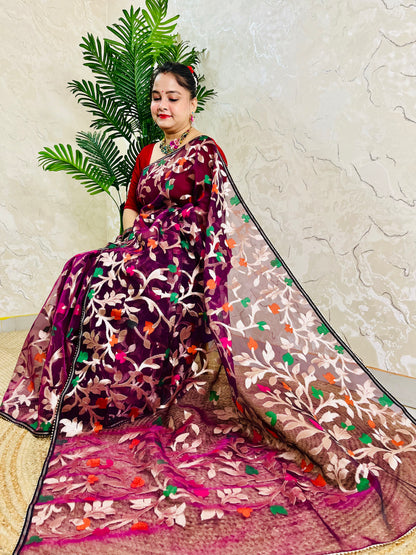 A woman wearing a traditional Indian saree with a deep purple body, brown border, and multicolor floral embroidery, posed next to a green plant against a textured beige wall.