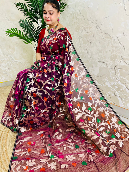 A woman wearing a purple and brown floral embroidered saree with a red blouse, seated indoors against a textured wall with a green plant in the background.