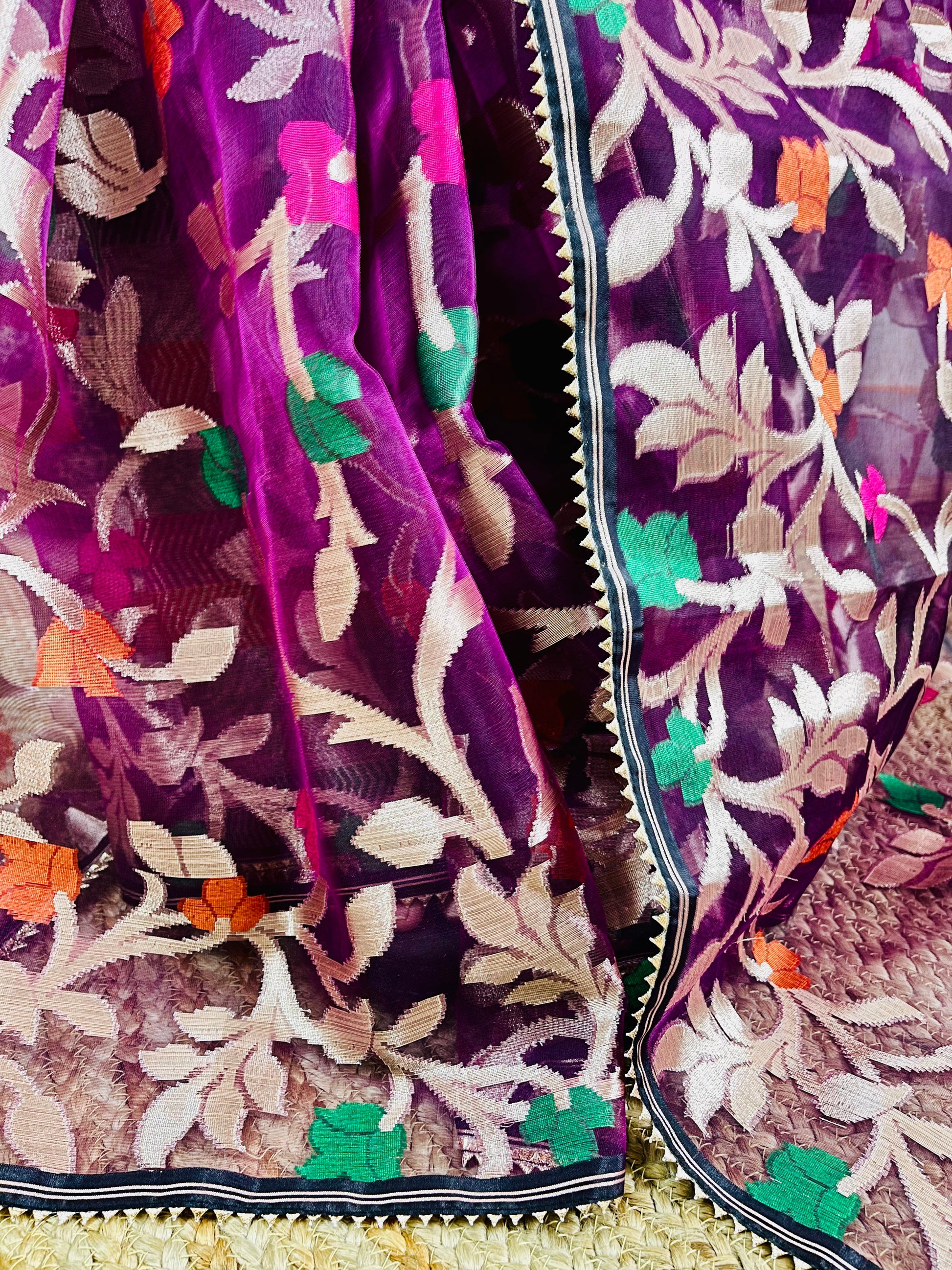 A close-up of a purple embroidered fabric, likely a saree or scarf, featuring intricate floral and leaf patterns in beige, green, orange, and pink on a sheer mesh base.