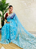 A woman wearing a light blue georgette saree with white floral embroidery and a matching dupatta, paired with a maroon blouse, seated on a woven mat in front of a textured beige wall.