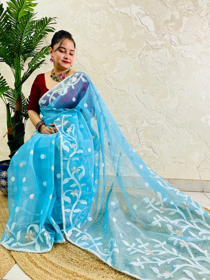 A woman wearing a light blue georgette saree with white floral embroidery and a matching dupatta, paired with a maroon blouse, seated on a woven mat in front of a textured beige wall.