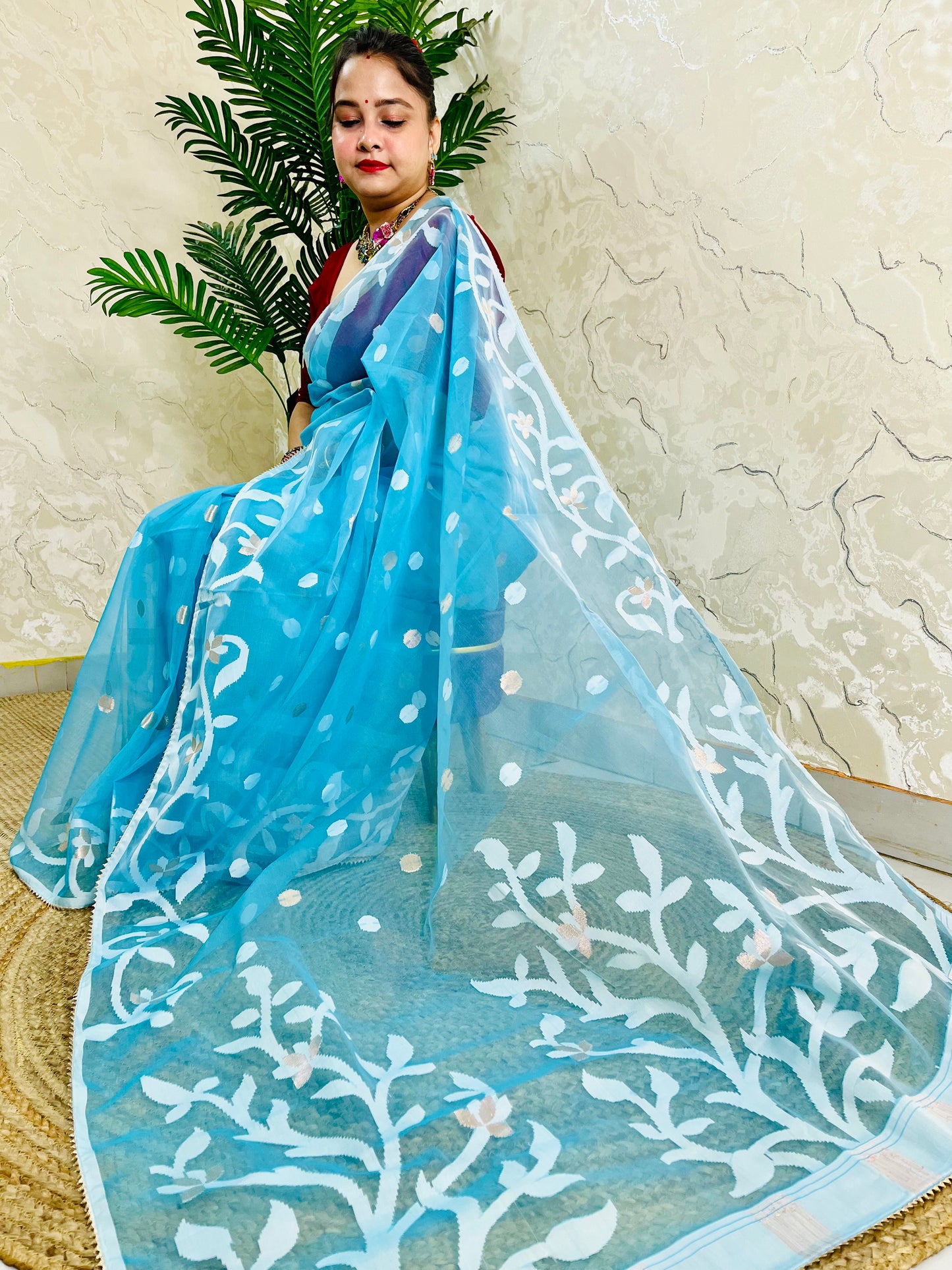 A woman wearing a light blue saree with white floral embroidery and white polka dots, posed in front of a textured beige wall with a green plant in the background.