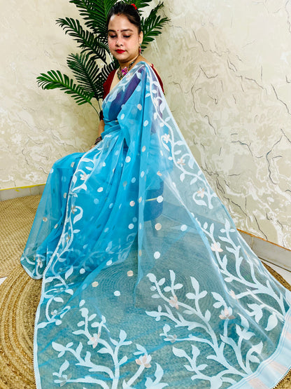A woman wearing a bright blue saree with white floral embroidery and a matching sheer dupatta, seated indoors against a textured beige wall with a green plant in the background.