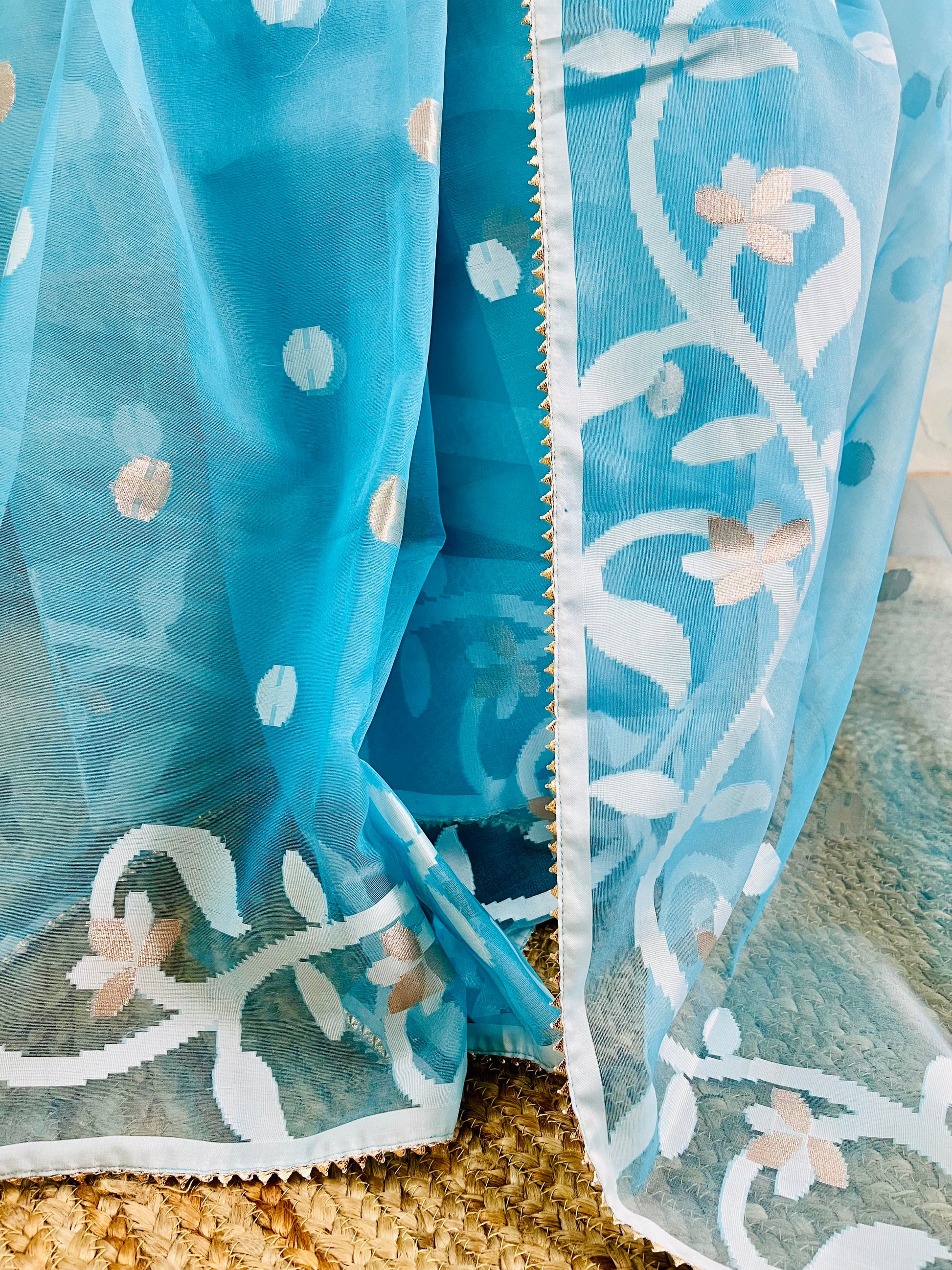 A close-up of a sheer turquoise blue fabric with white floral embroidery and small metallic gold dots, draped over a textured beige woven surface.