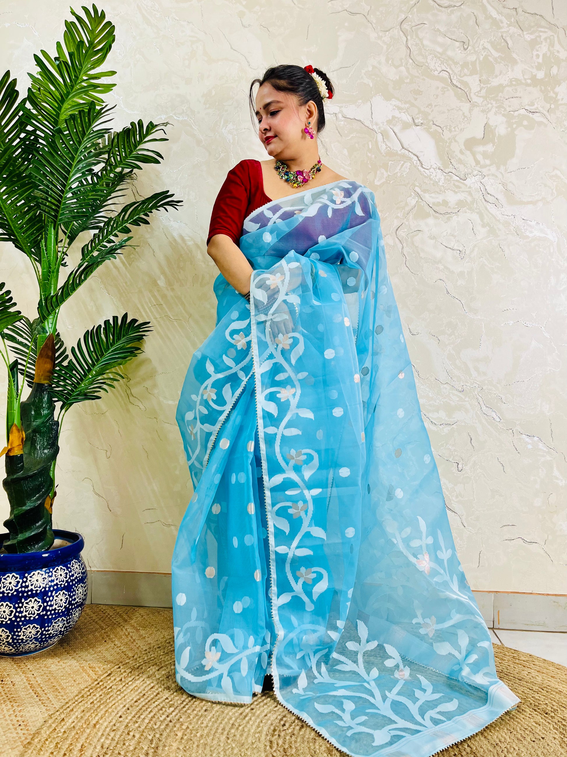 A woman wearing a light blue saree with white floral patterns and a red sleeveless blouse, standing indoors next to a potted plant against a textured beige wall.