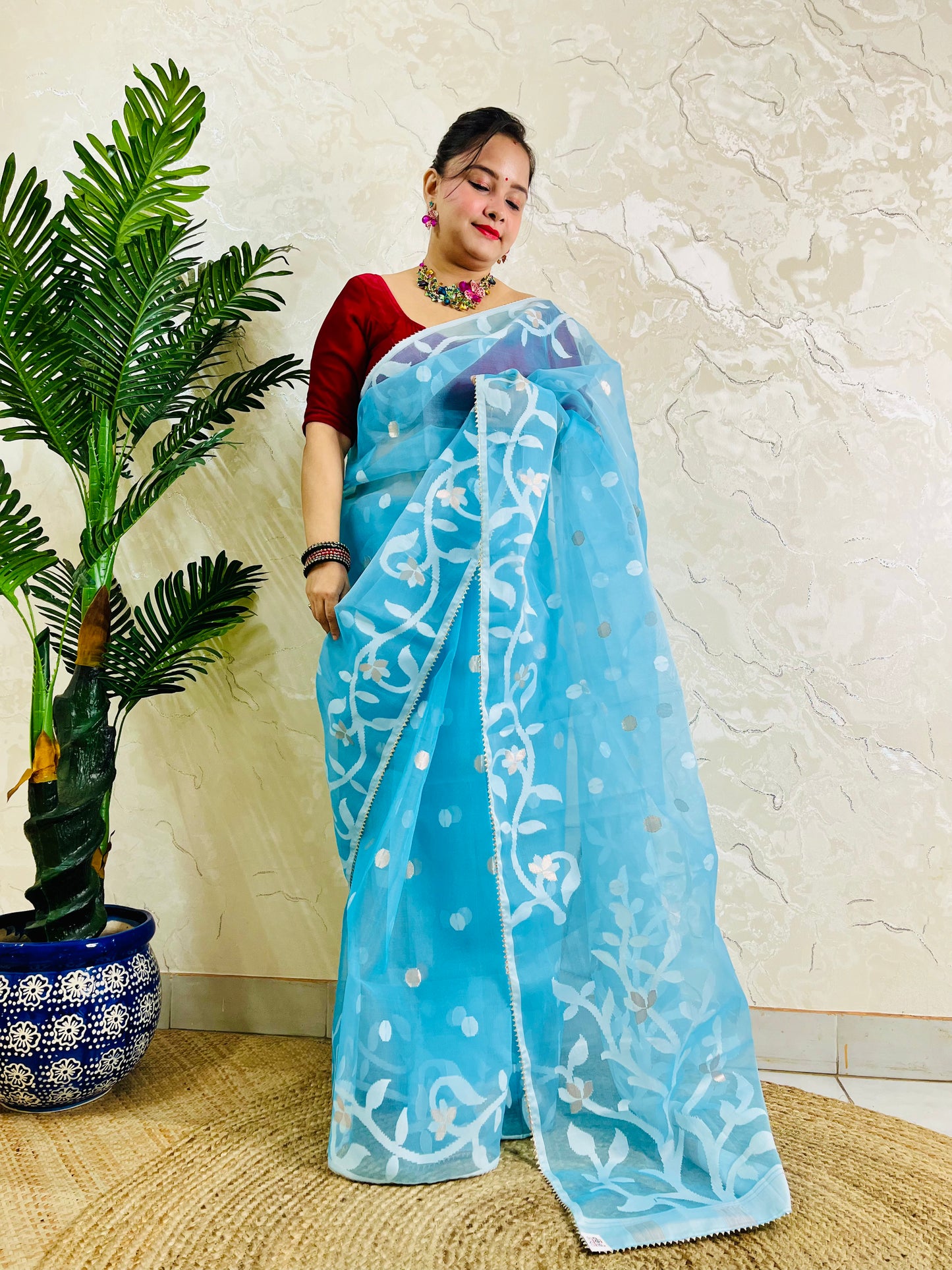 A woman wearing a light blue saree with white floral embroidery and a red blouse, standing indoors next to a potted palm tree against a textured beige wall.