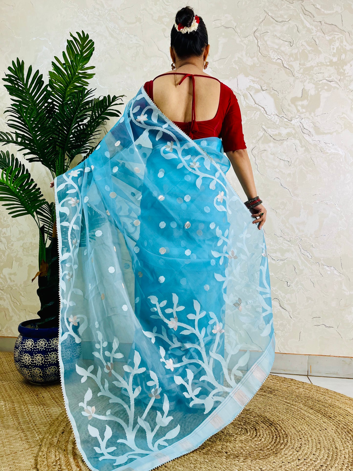 A woman shown from behind wearing a light blue saree with white floral embroidery and a red blouse, standing indoors against a textured beige wall with a potted plant on the left.