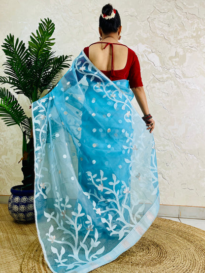 A woman shown from behind wearing a light blue saree with white floral embroidery and a red blouse, standing indoors against a textured beige wall with a potted plant on the left.