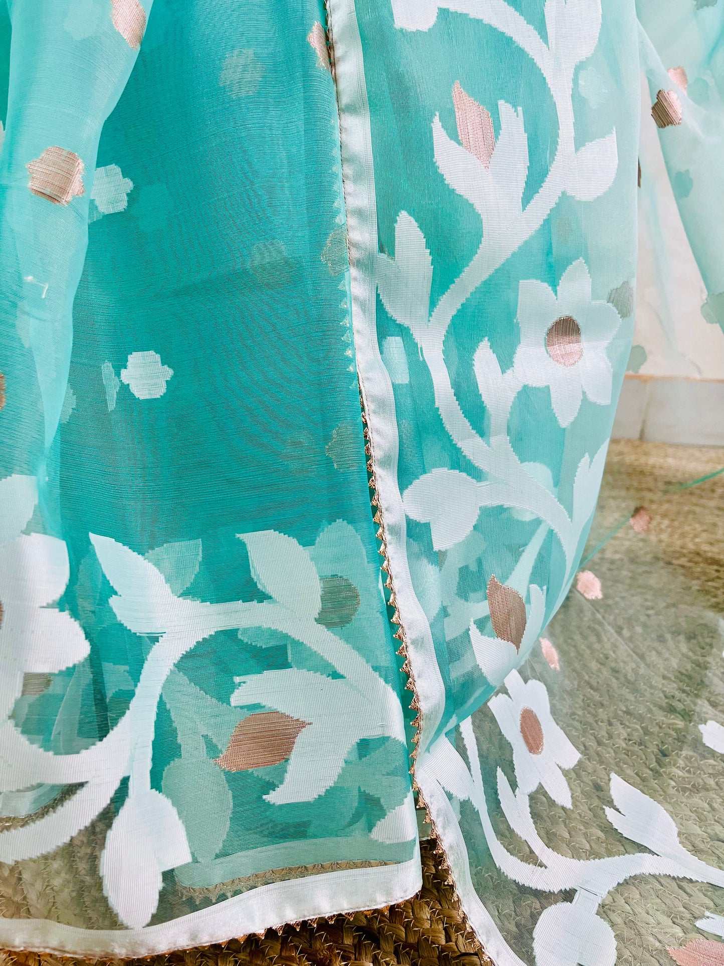 A close-up of a teal and white embroidered fabric, likely a dupatta or sari, featuring floral patterns with gold foil accents and a white border.