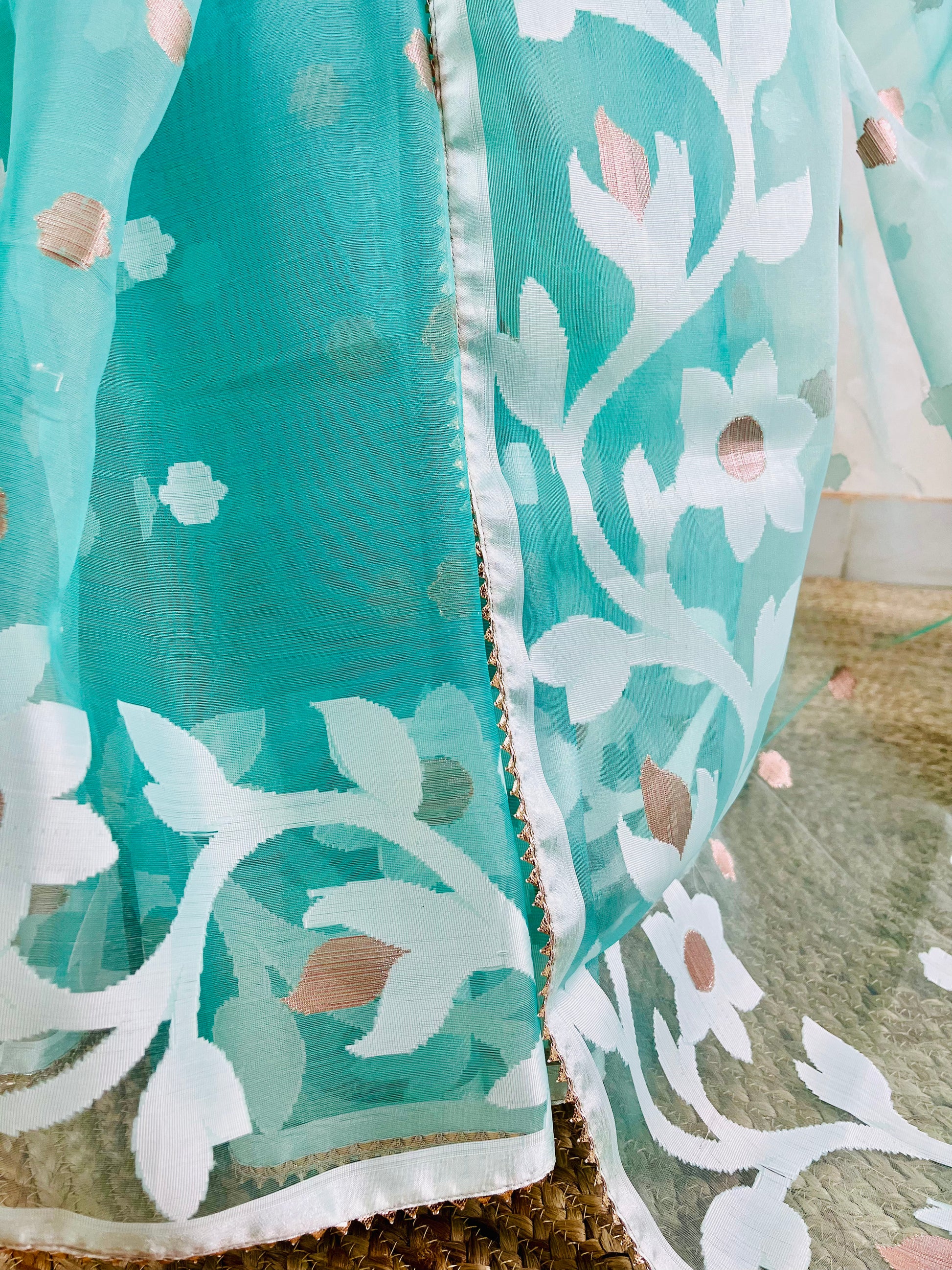 A close-up of a teal and white embroidered fabric, likely a dupatta or sari, featuring floral patterns with gold foil accents and a white border.